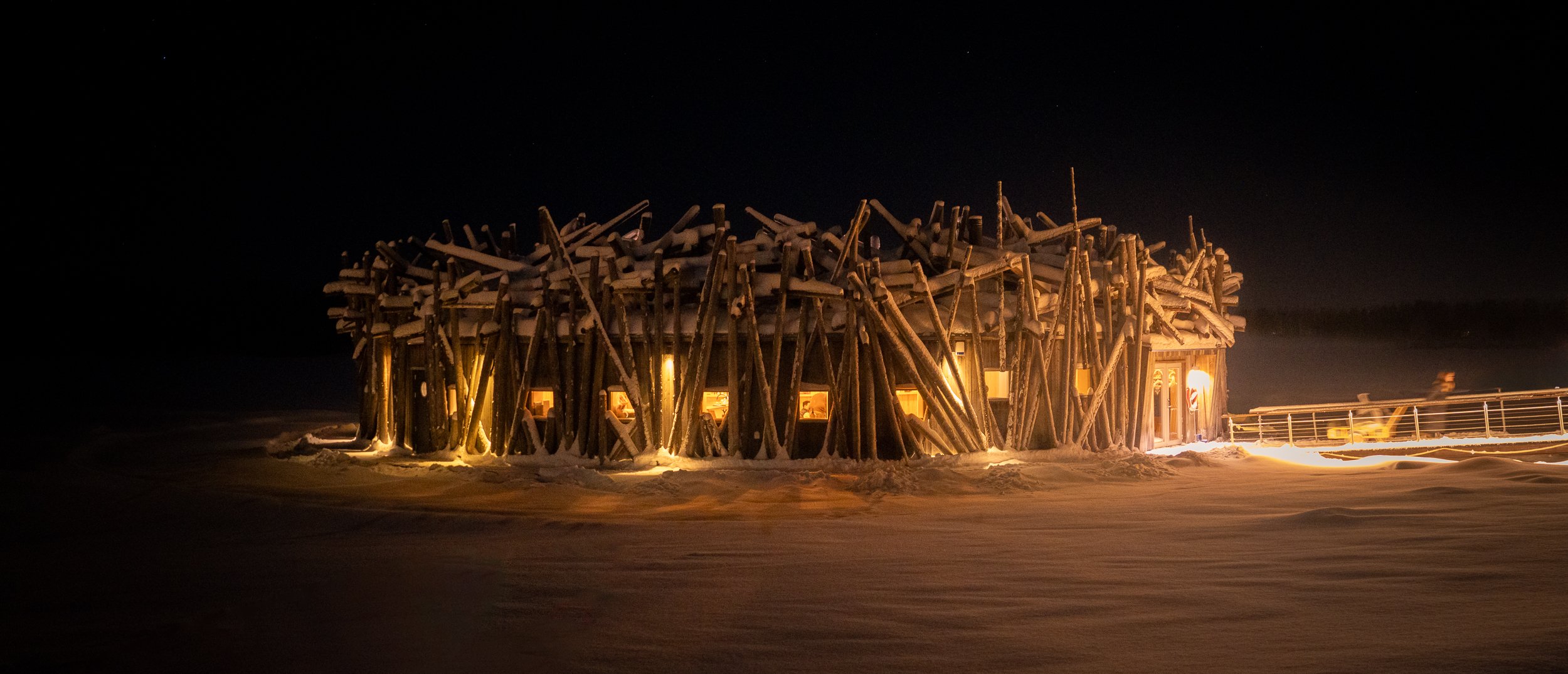 A large snow-covered wooden structure lit from within at night, with a wooden pier extending to the right.