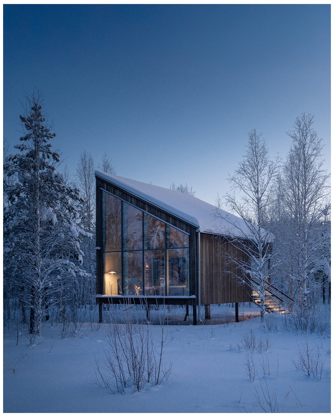 A modern house with large glass windows, surrounded by snow-covered trees and landscape, with lights glowing inside during dusk or early evening.