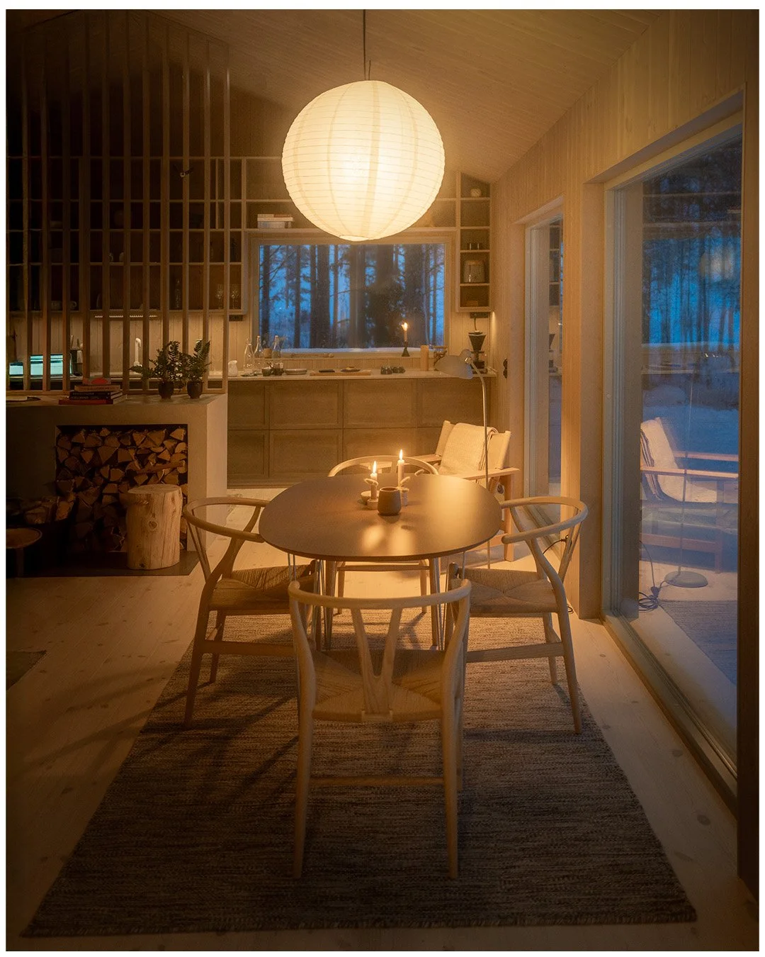 A cozy dining area illuminated by a large paper lantern pendant light, with a round wooden table, candles, and four chairs, set in a wooden interior with large sliding glass doors and a view of a wooded yard.