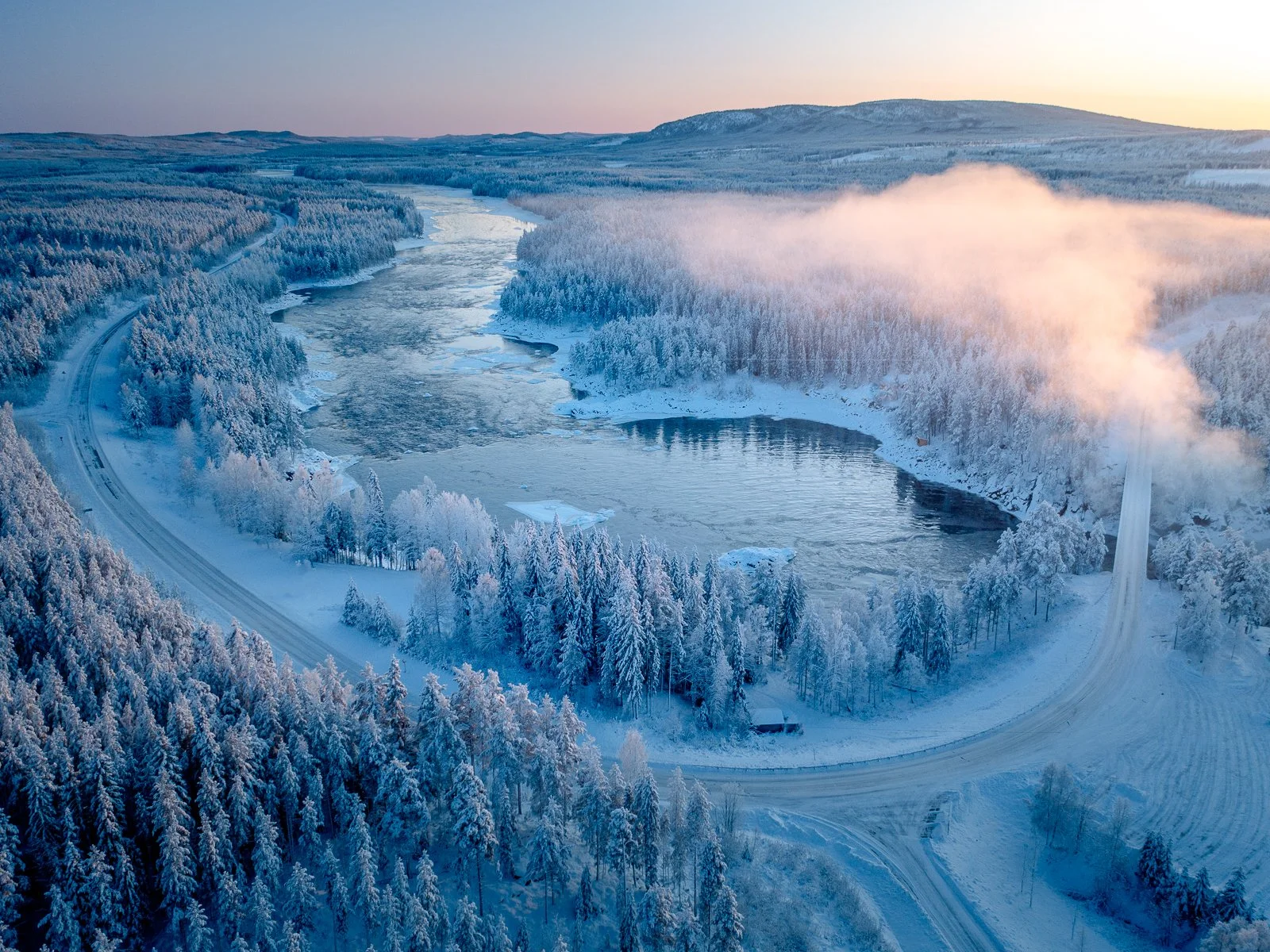 A snowy landscape with a partially frozen river winding through a forest covered in snow. There is a railway track on the left side and a chimney emitting smoke on the right side with a sunset in the background.
