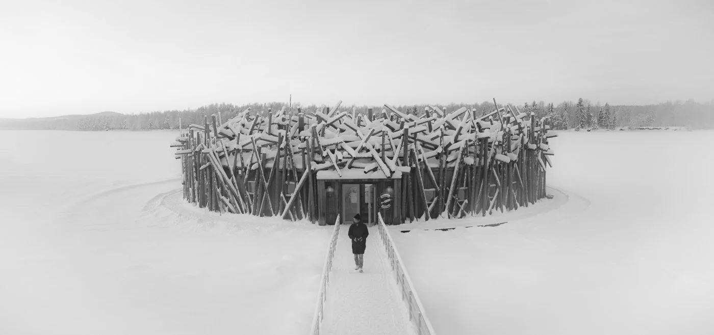 A person walking on a snow-covered walkway toward a round wooden building with a roof made of tilted logs, set in a snowy landscape.