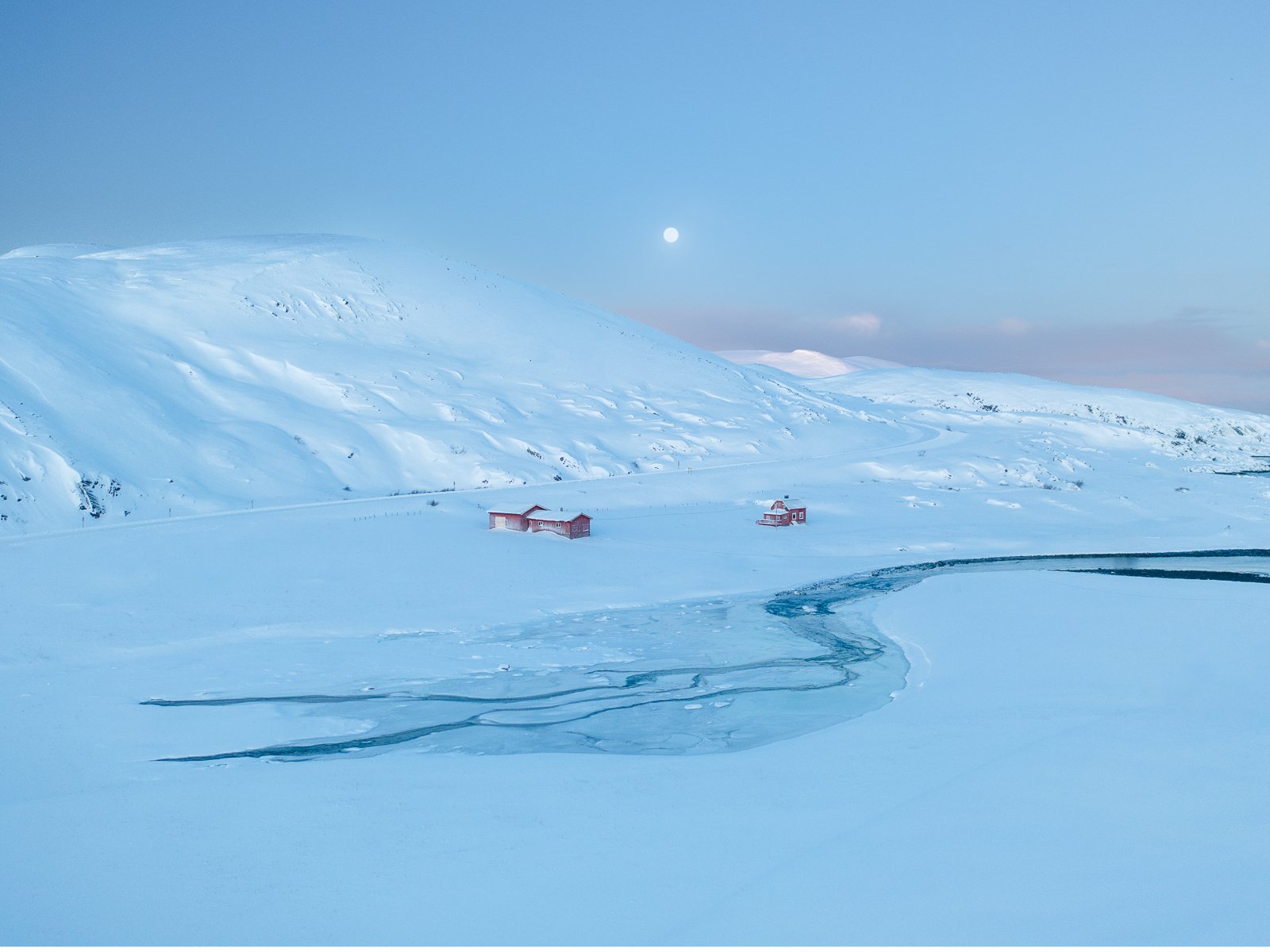 Snow-covered landscape with two small red buildings, a winding frozen river, a hill in the background, and a full moon in the sky.