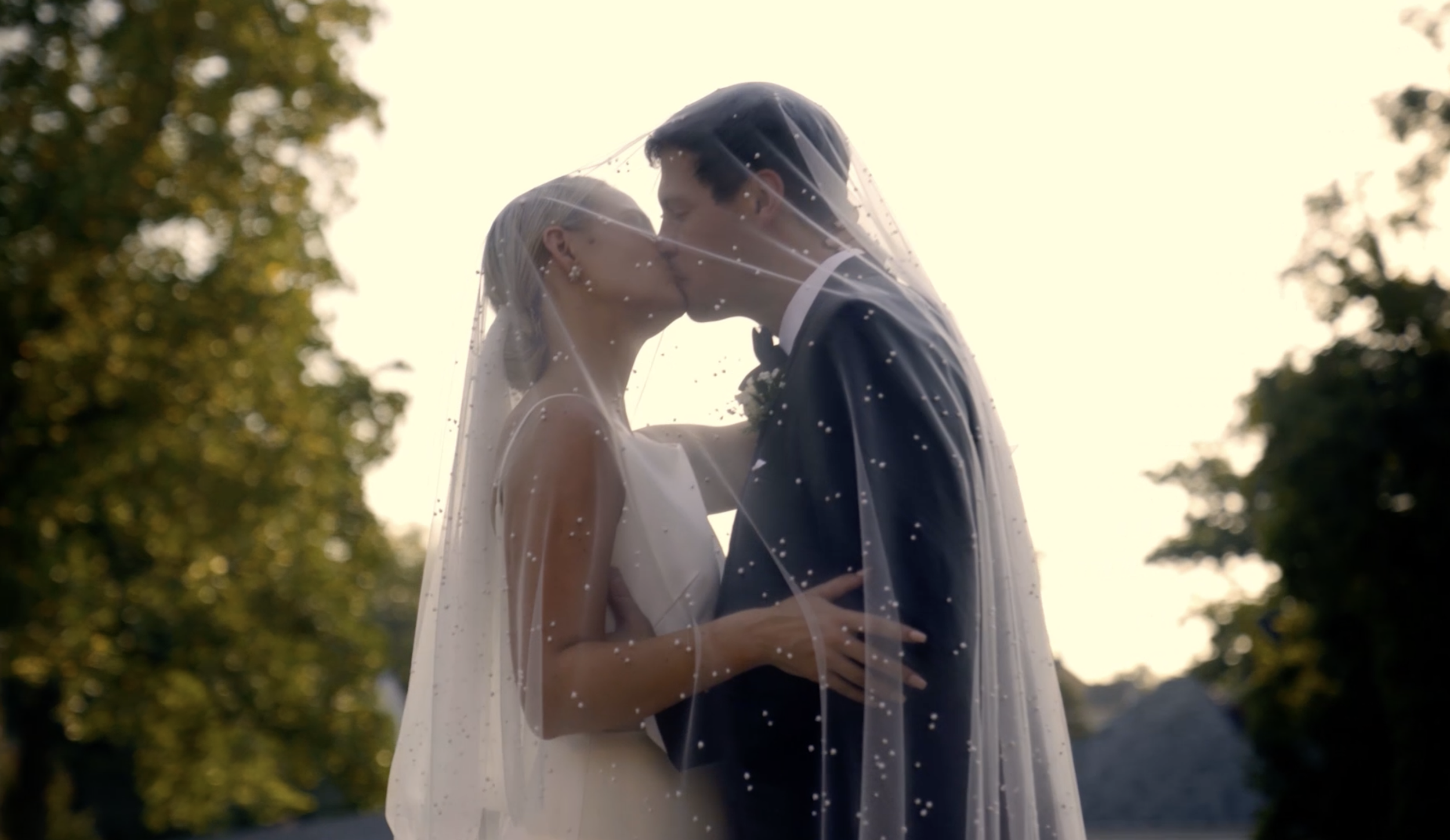 A bride and groom kissing under a veil outdoors during sunset, with trees in the background.