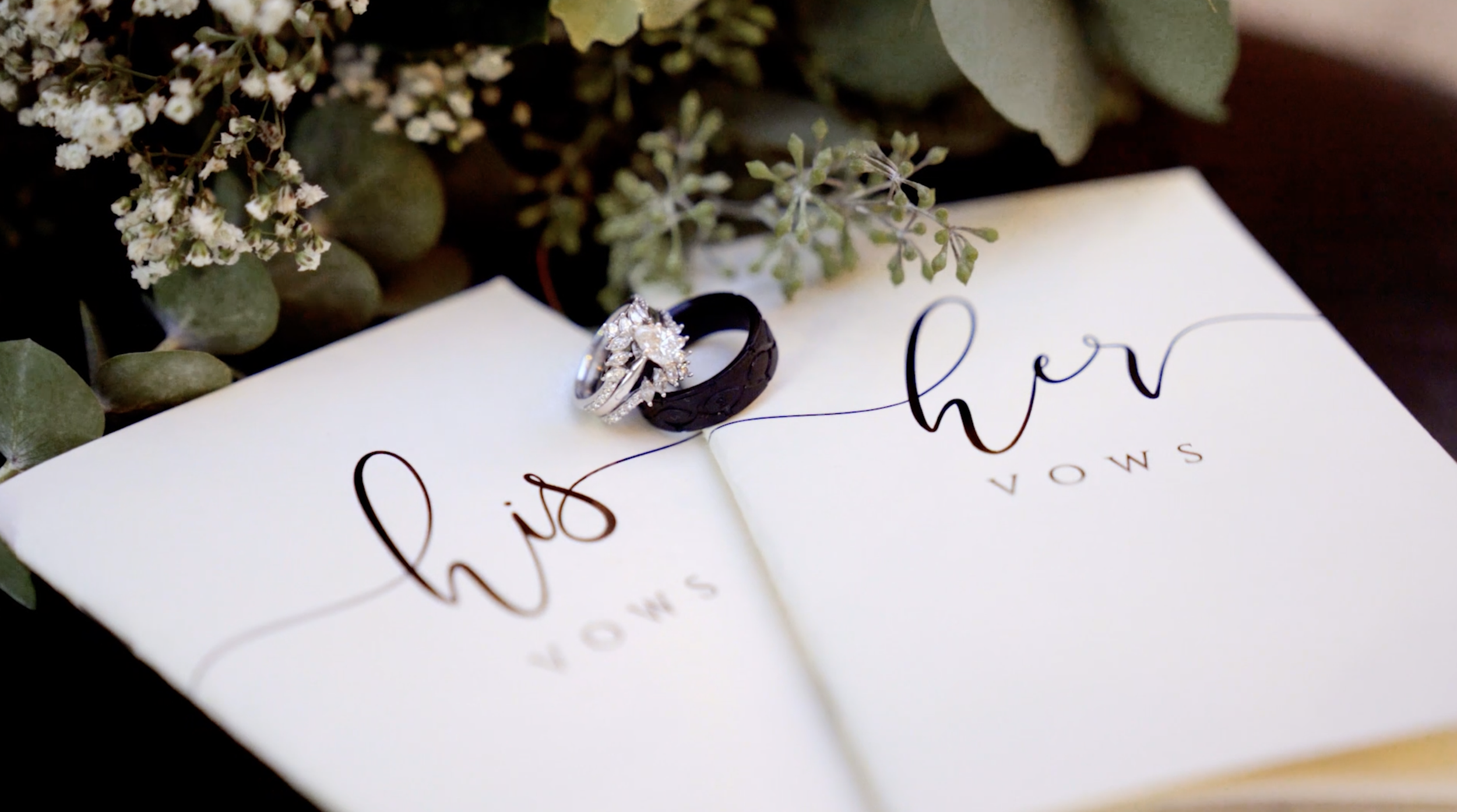 A wedding vow booklet with rings placed on top, surrounded by greenery and white flowers.