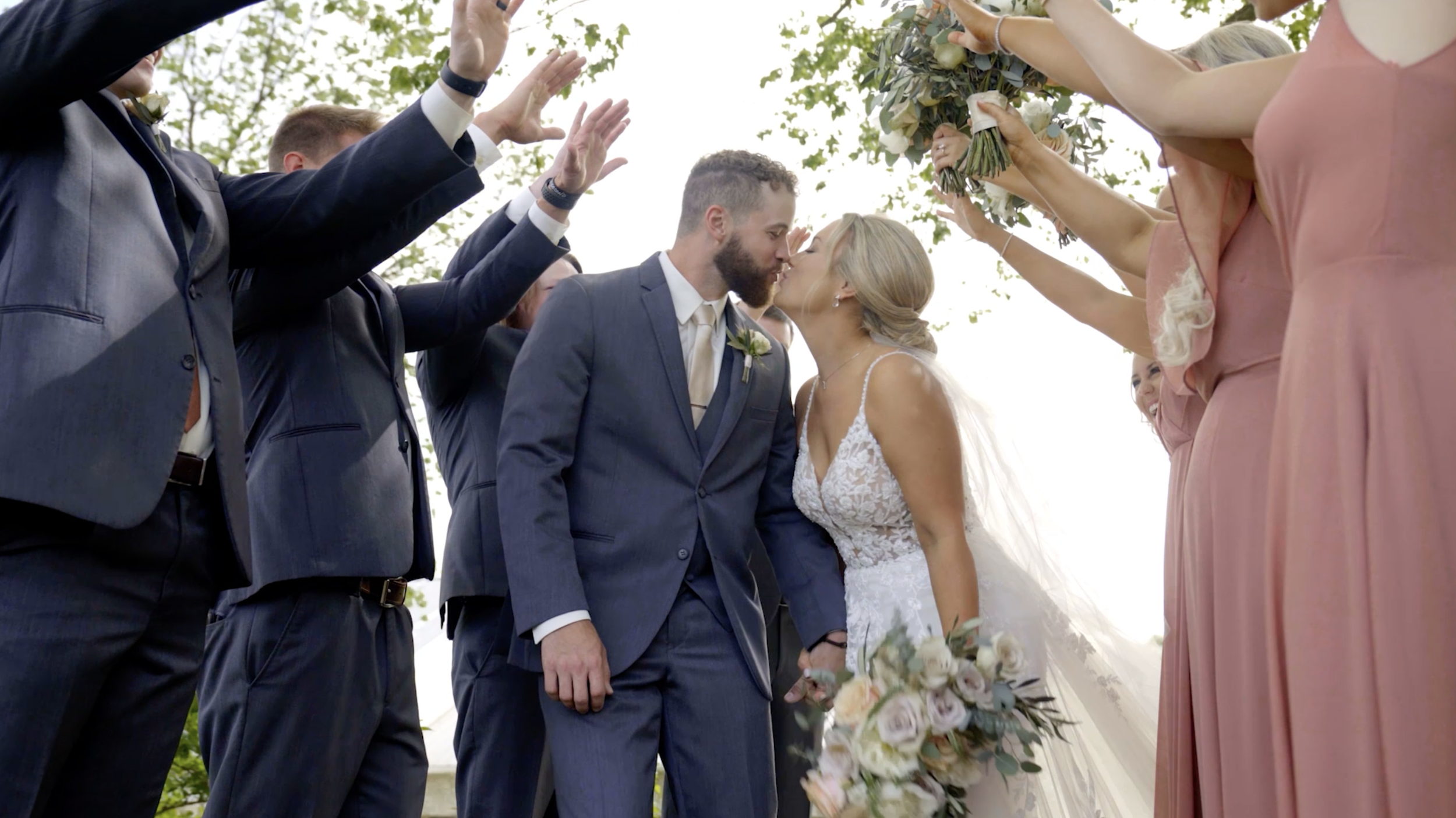 Bride and groom kissing under a floral arch while friends and family raise their hands in celebration, outdoors during a wedding ceremony.