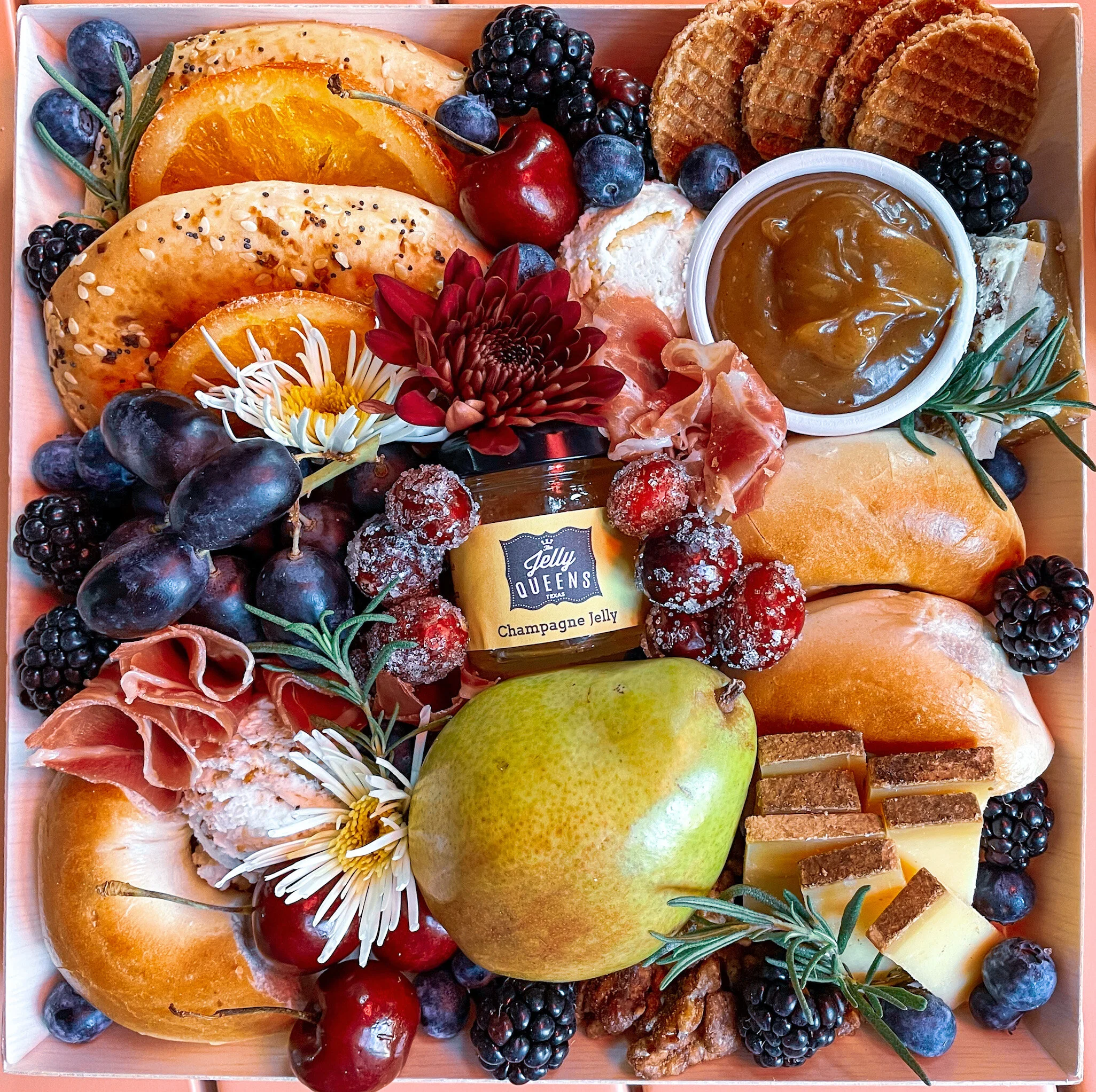 Charcuterie board with bagels, fruit, prosciutto, cheese, champagne jelly, caramel dip, and flowers.