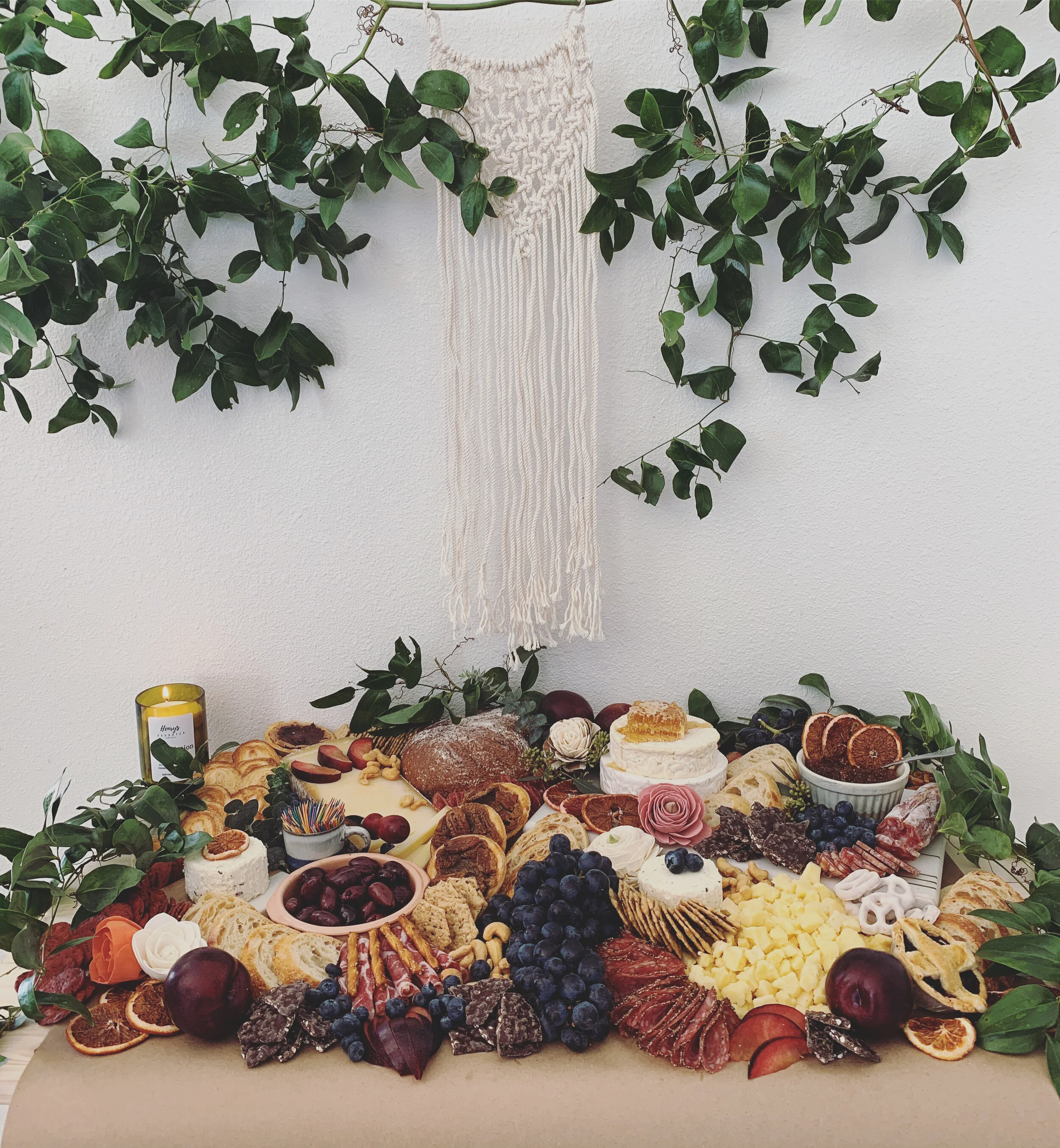 A rustic charcuterie board with assorted cheeses, meats, fruits, nuts, and crackers, surrounded by greenery and a macrame wall hanging.