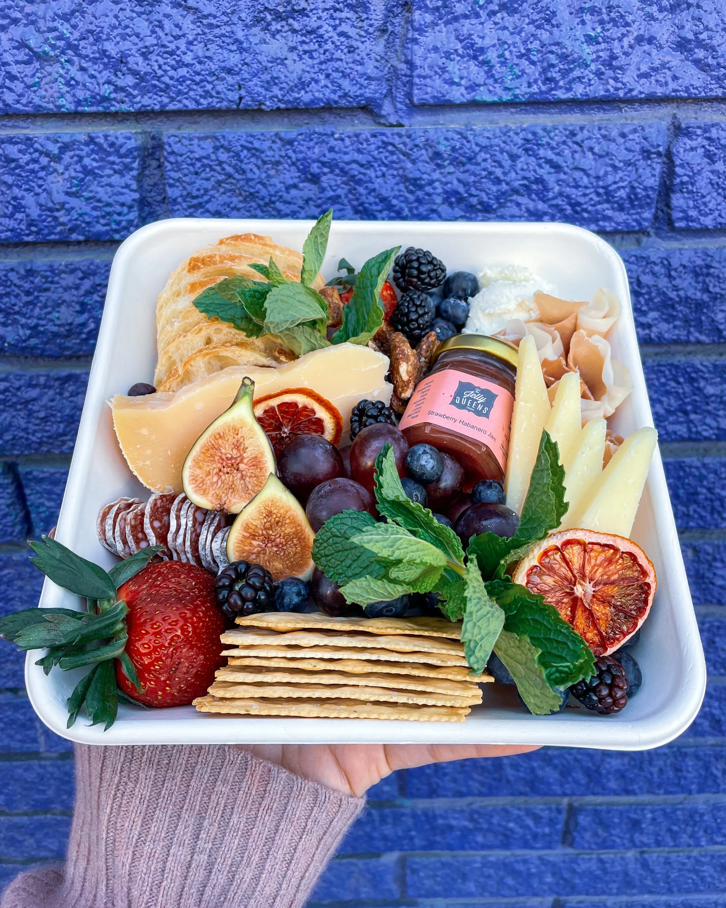 A charcuterie plate containing various cheeses, bread slices, figs, grapes, strawberries, blackberries, blueberries, crackers, mint leaves, and a small jar of strawberry habanero jam, set against a blue brick wall.