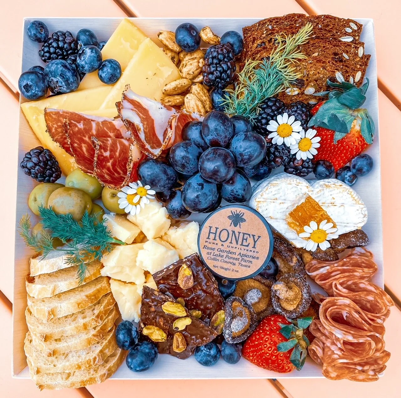 Charcuterie board with cheese, salami, grapes, berries, nuts, crackers, honey, and floral garnishes.