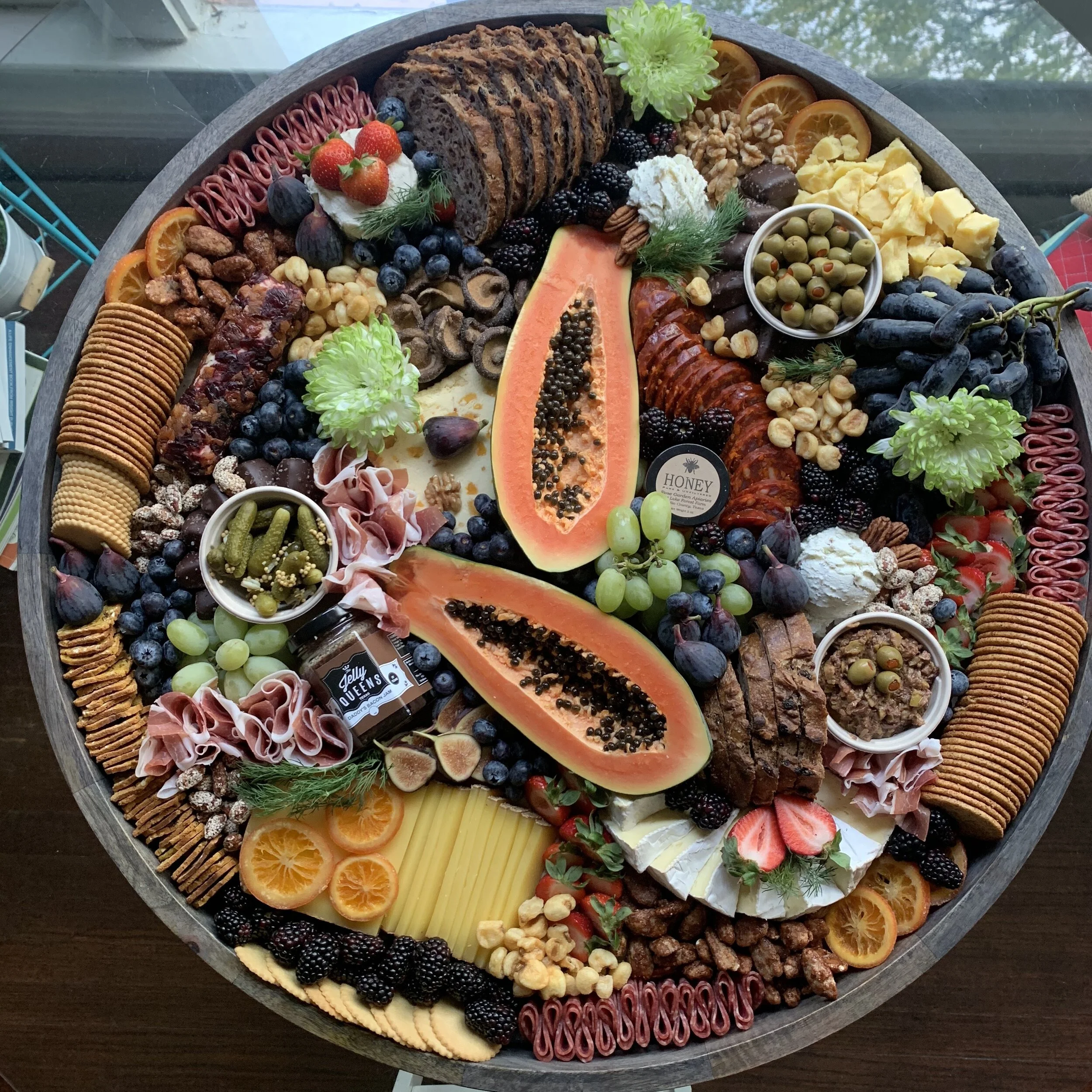 A large charcuterie board featuring a variety of foods including slices of bread, crackers, papaya, grapes, strawberries, olives, cheese, meats, nuts, and figs, arranged artfully with decorative flowers.