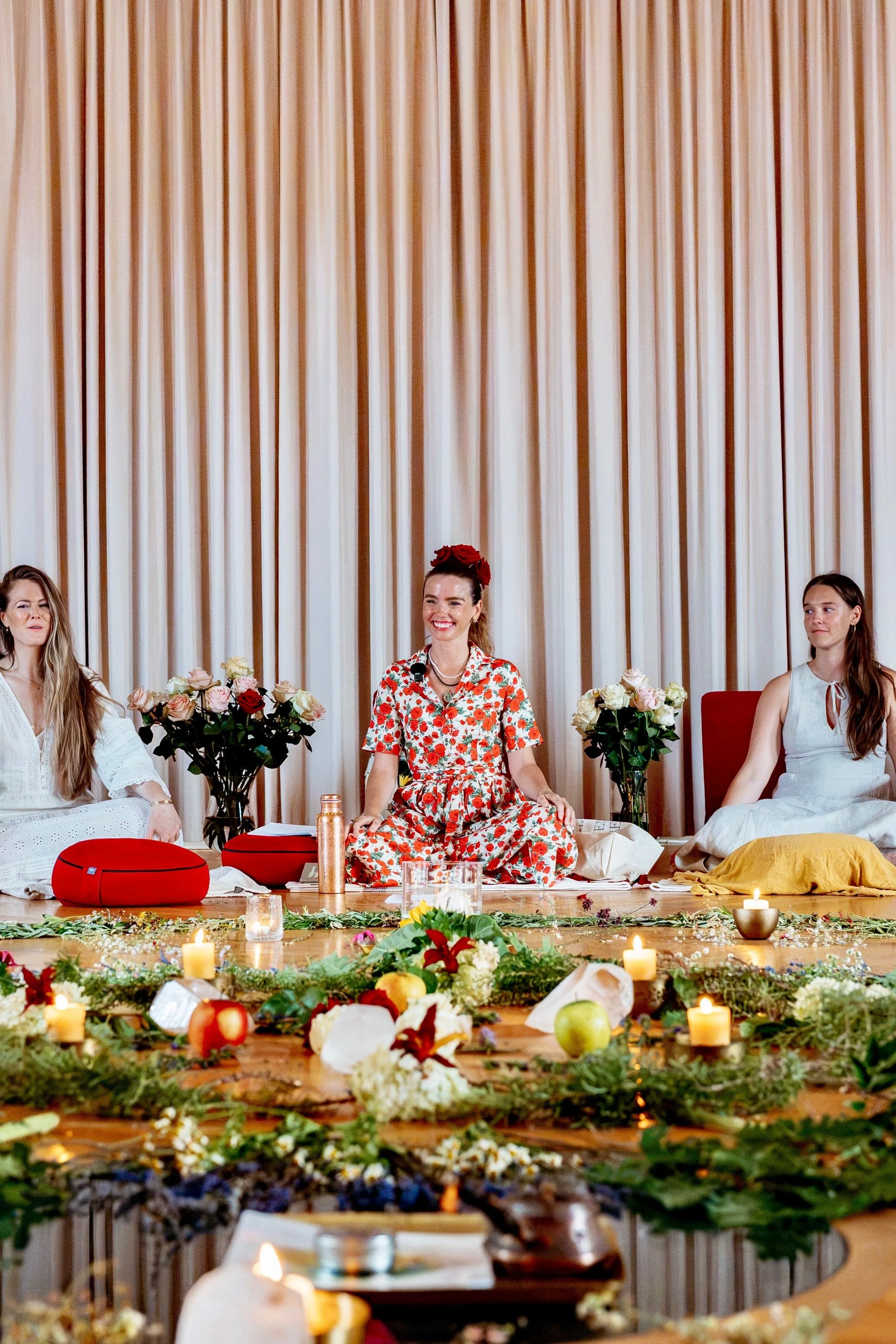 A group of women participate in a celebratory or ritualistic activity outdoors, with one woman in a blue dress smiling at the center and others raising their hands in unison behind her, on a grassy area near a building with large glass windows.