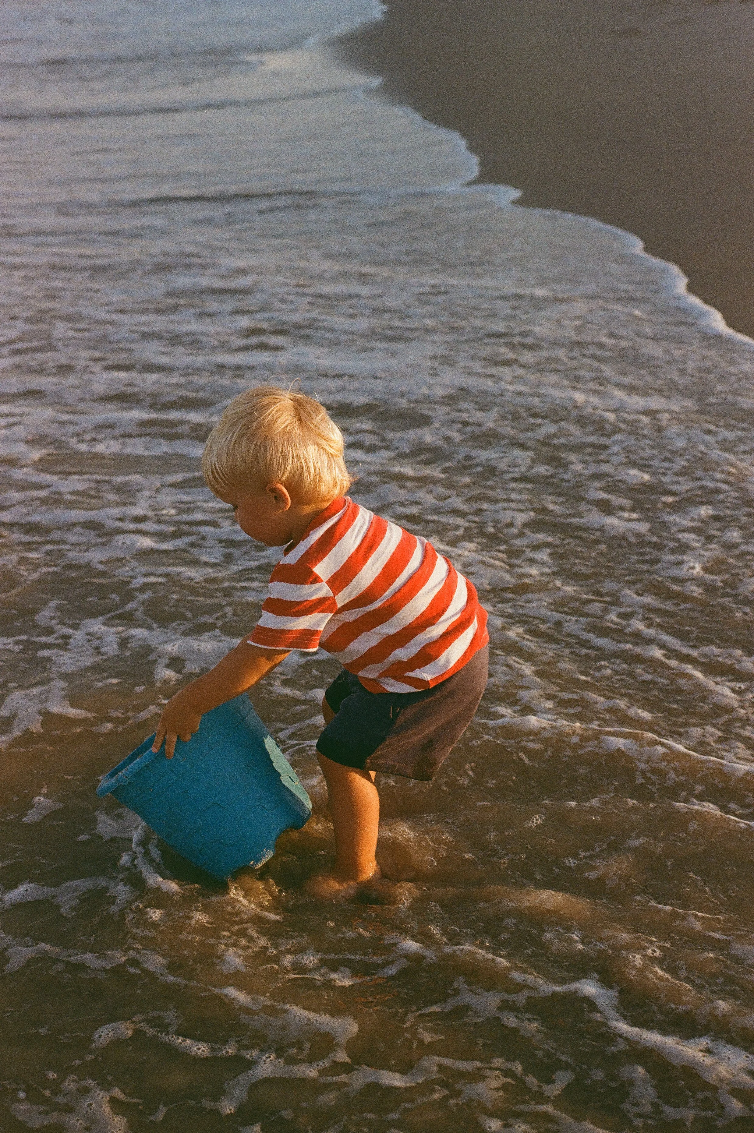 A young boy playing at the beach, crouched down and holding a blue bucket in the shallow water near the shore during sunset.