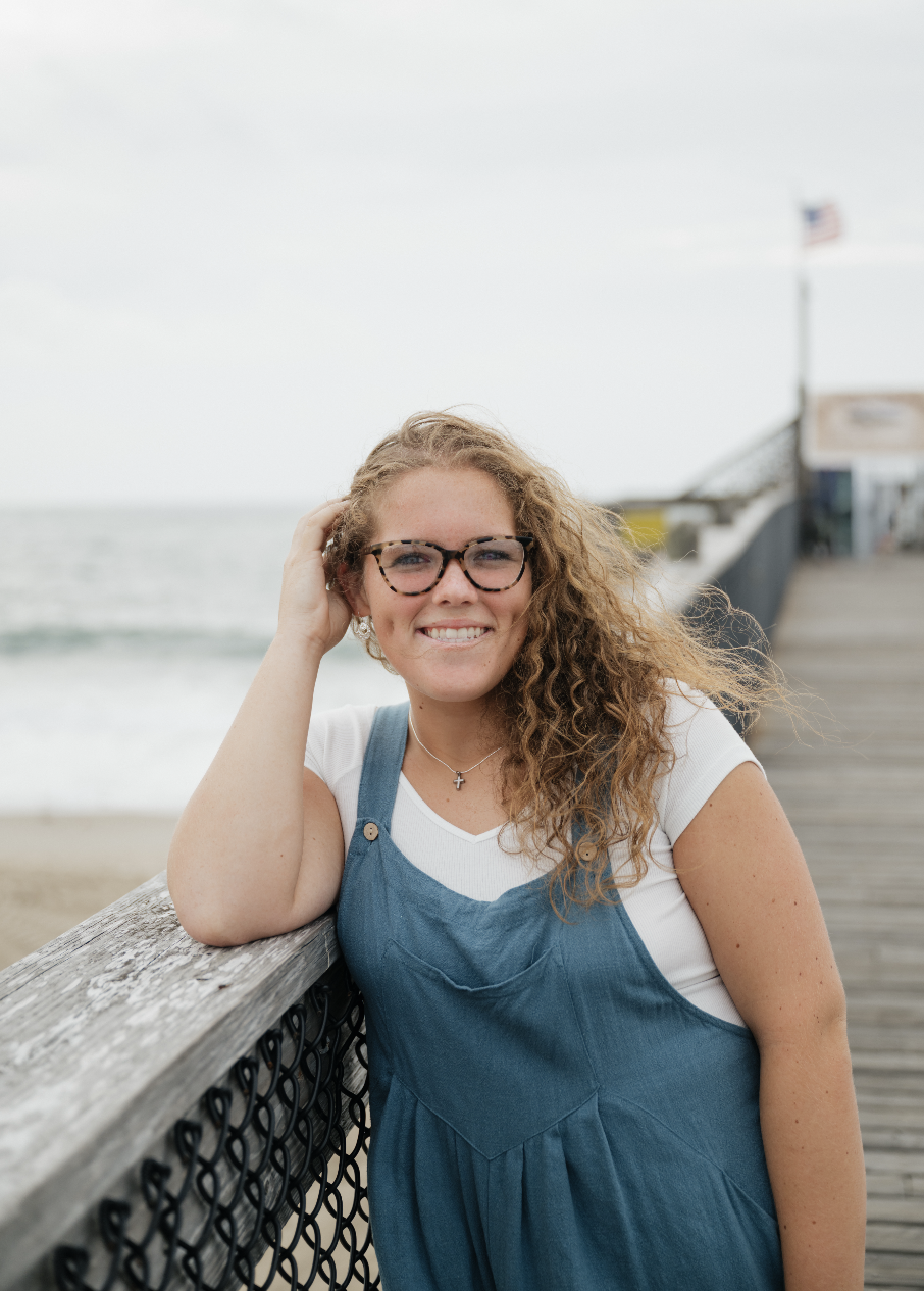 A smiling young woman with curly hair and glasses leaning on a beachside railing, in front of the ocean and a wooden pier