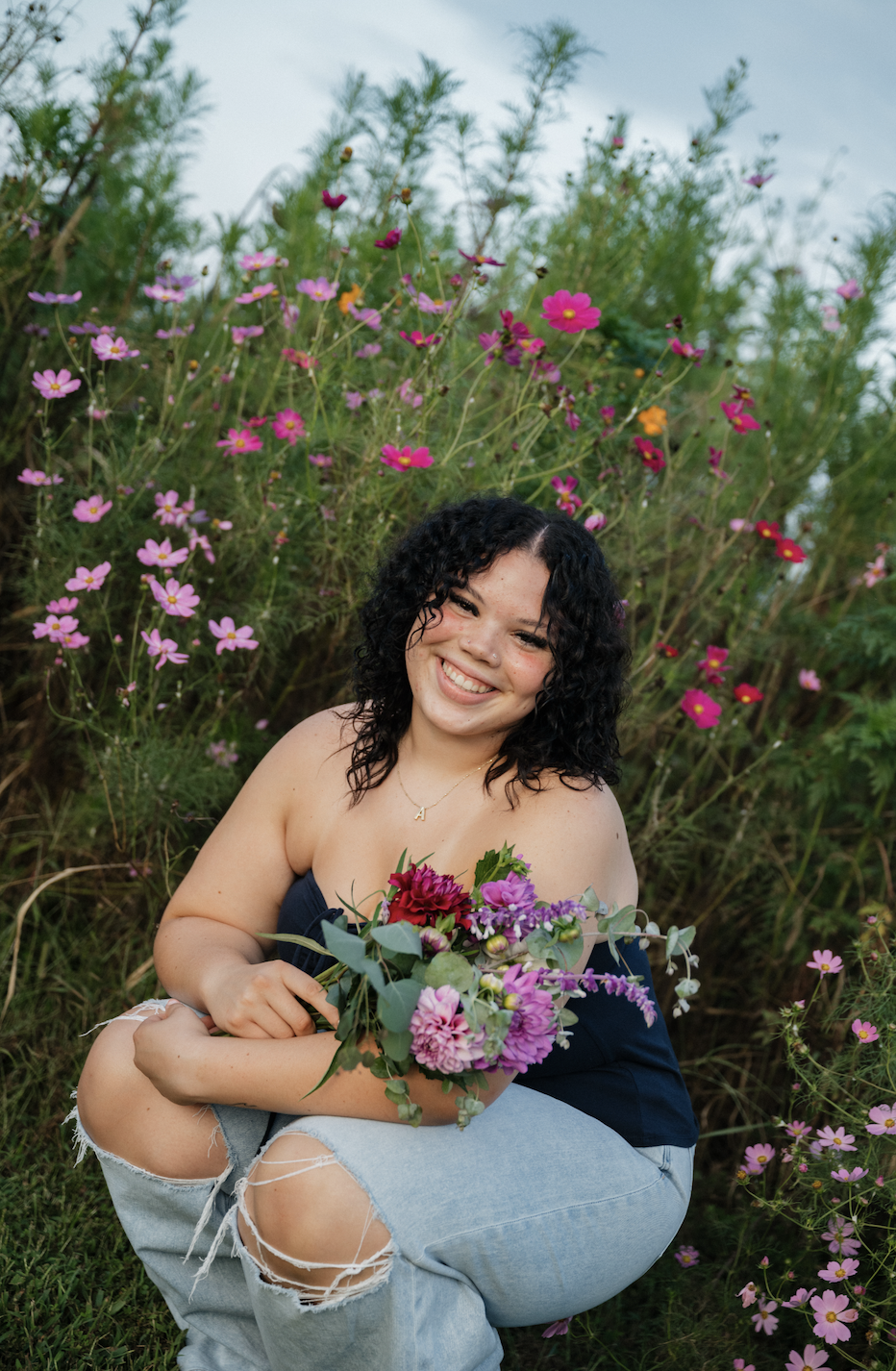 A young woman with curly black hair and a big smile, sitting outdoors in front of flowering plants, holding a bouquet of purple, pink, and red flowers.