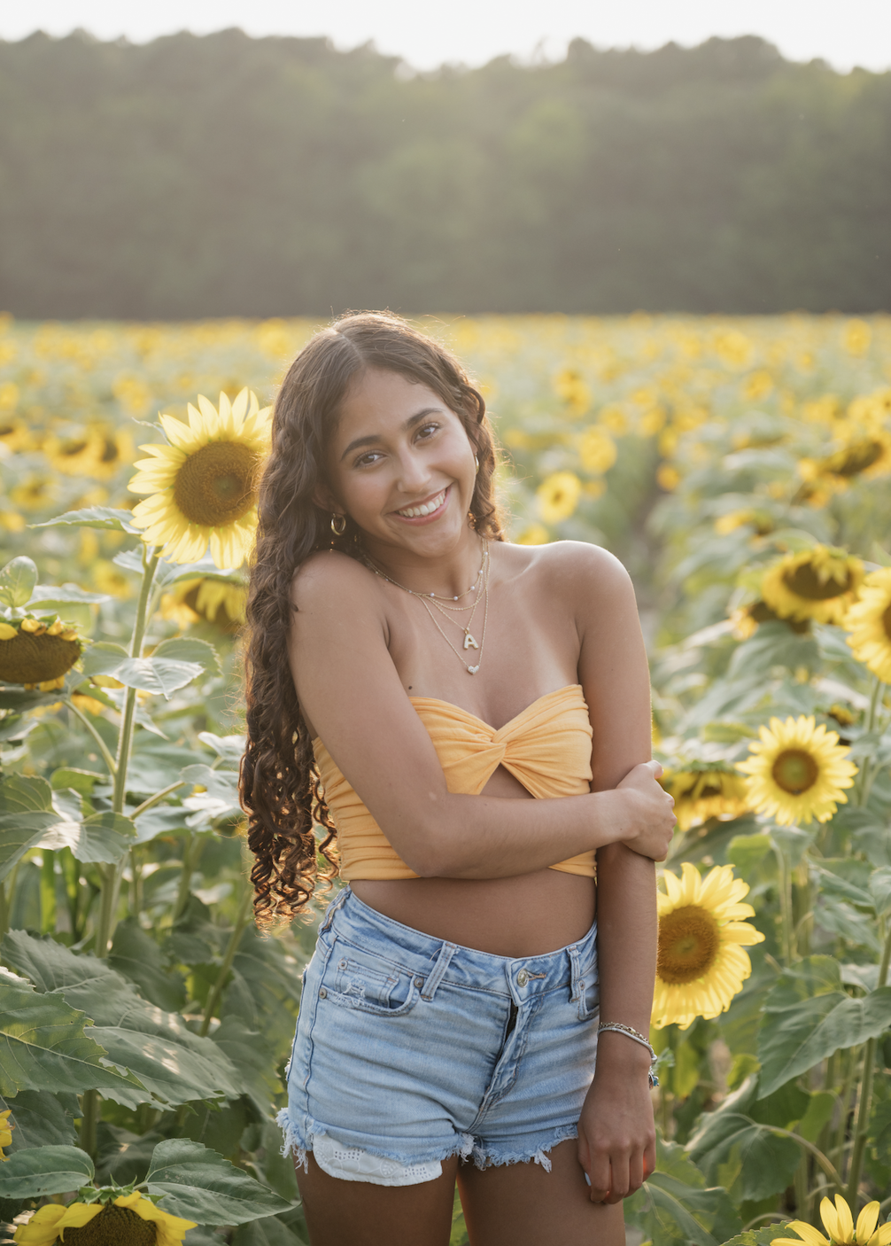 A young woman with long curly hair smiling in a sunflower field during sunset, wearing a yellow strapless bandeau top and denim shorts.