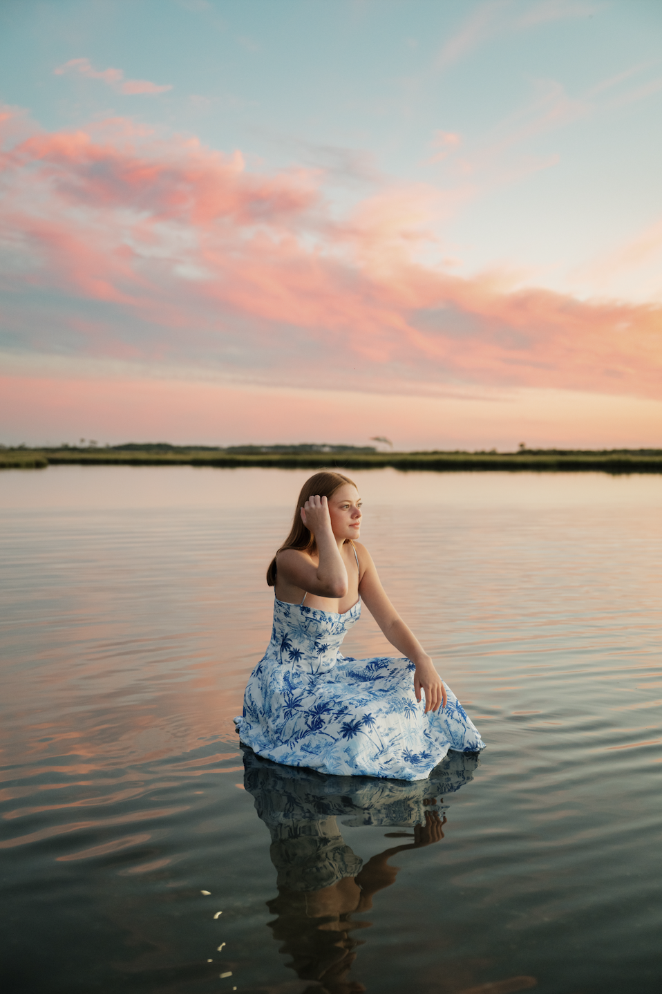 A young woman sitting in shallow water at sunset, wearing a blue and white dress with a palm tree pattern, with pink and orange clouds in the sky reflected on the water.