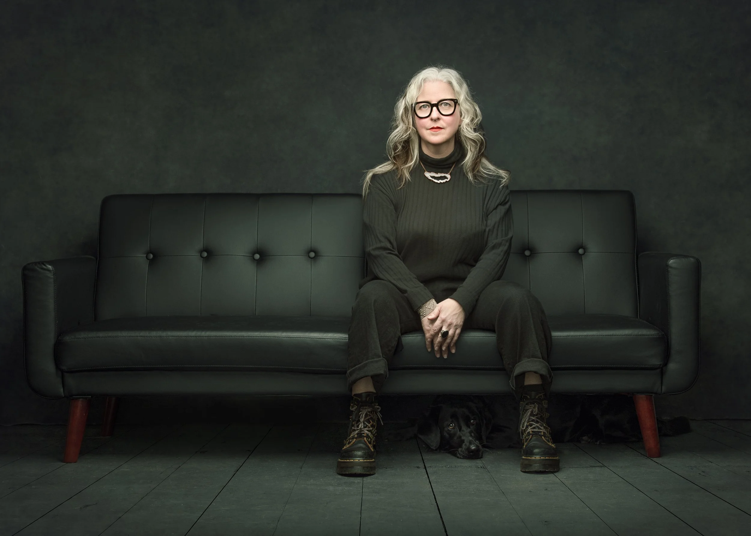 Woman sitting on a dark green couch with a dog peeking out underneath, wearing glasses and dark clothing, with a moody, dimly lit background.