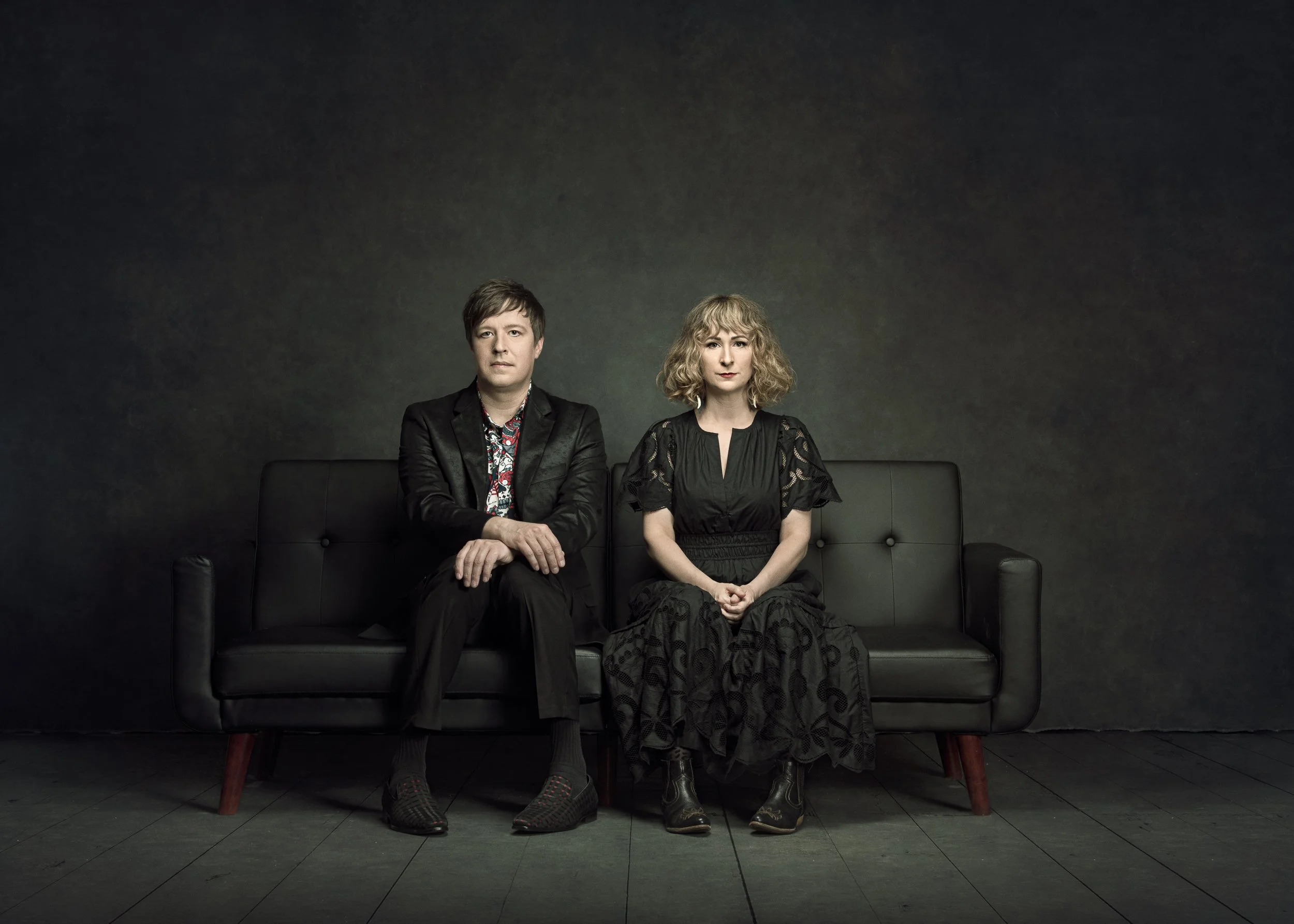 Husband and wife sitting on a black sofa against a dark backdrop.