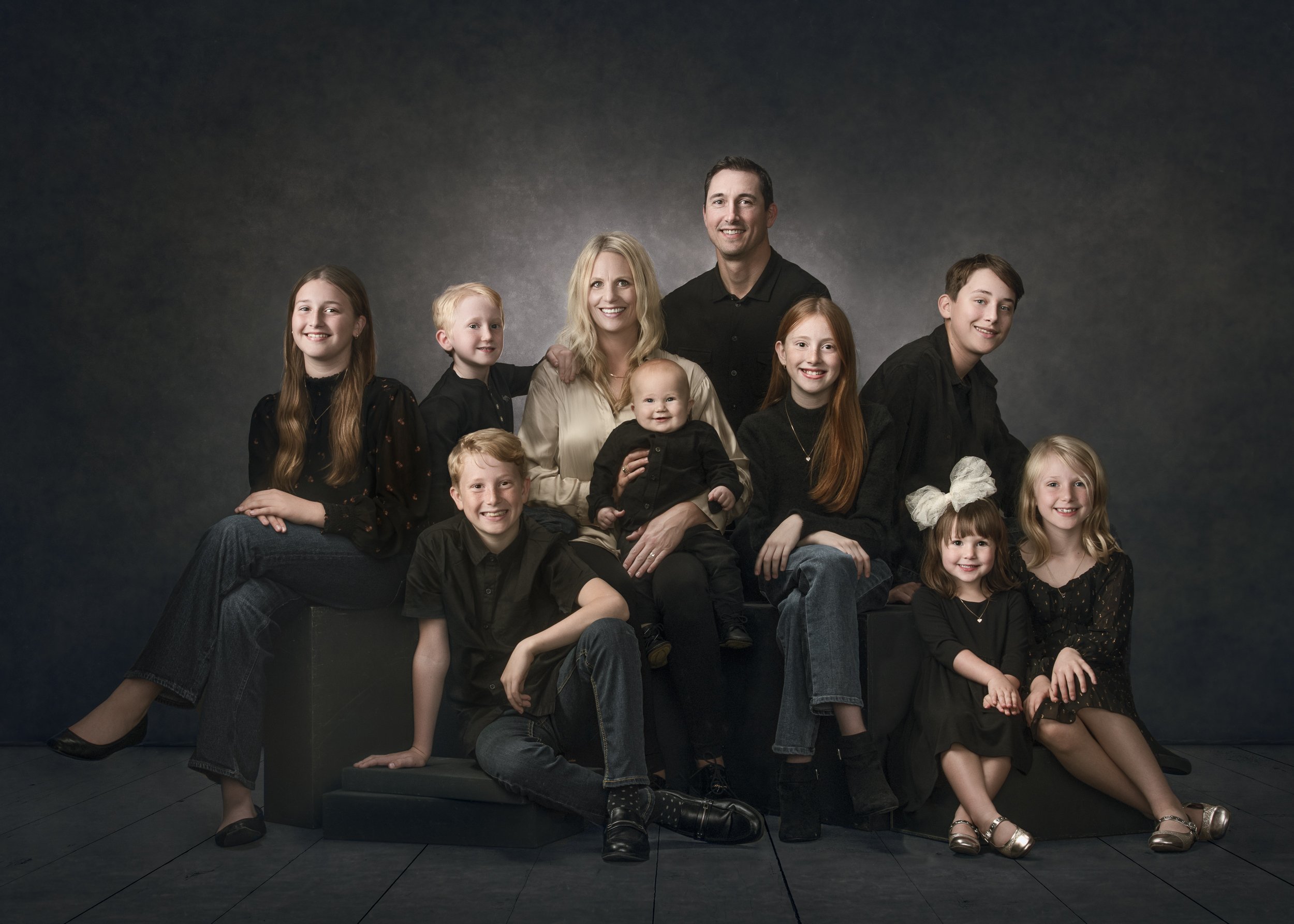 Family portrait with nine members in dark clothing against a studio background.