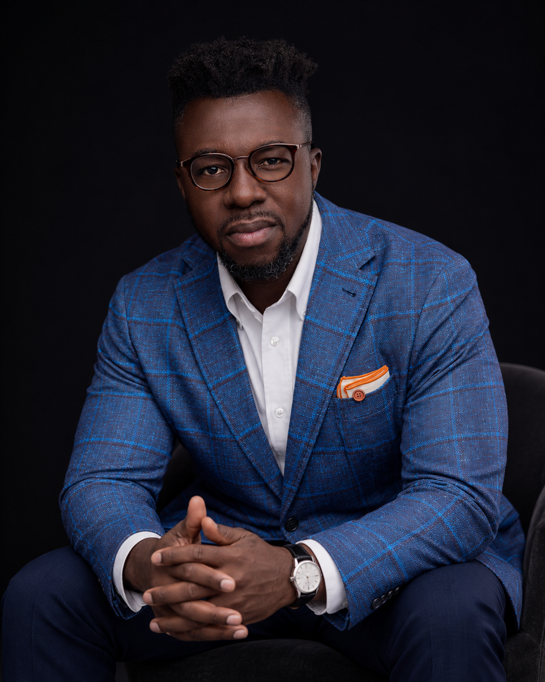 A man in a blue plaid blazer with glasses, white shirt, and a watch, sitting against a black background, looking directly at the camera with hands clasped.