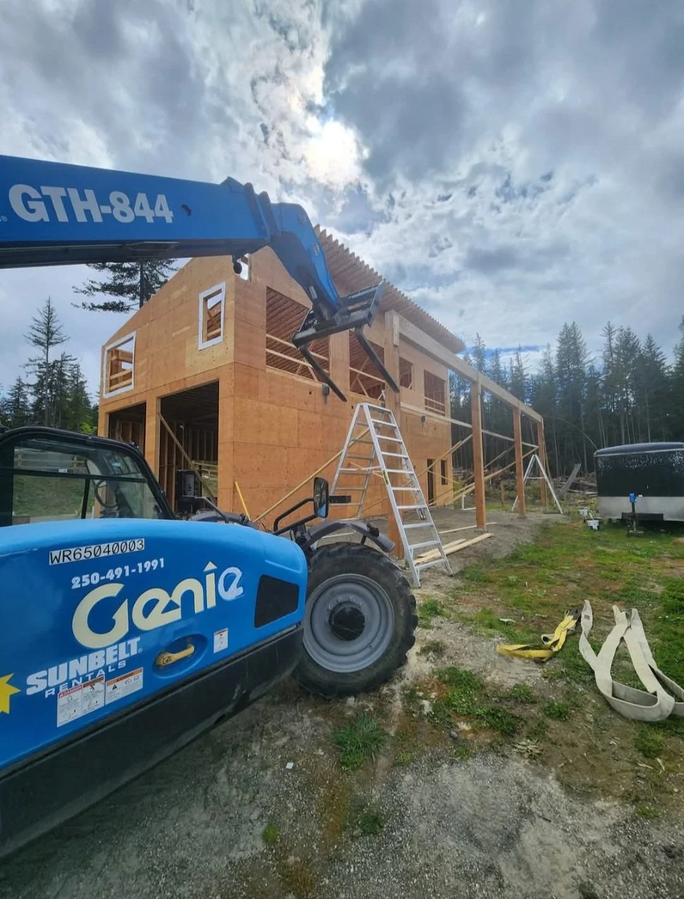 Construction site with a partly built wooden house, construction equipment, a large blue crane, a ladder, and a trailer, with trees and a cloudy sky in the background.