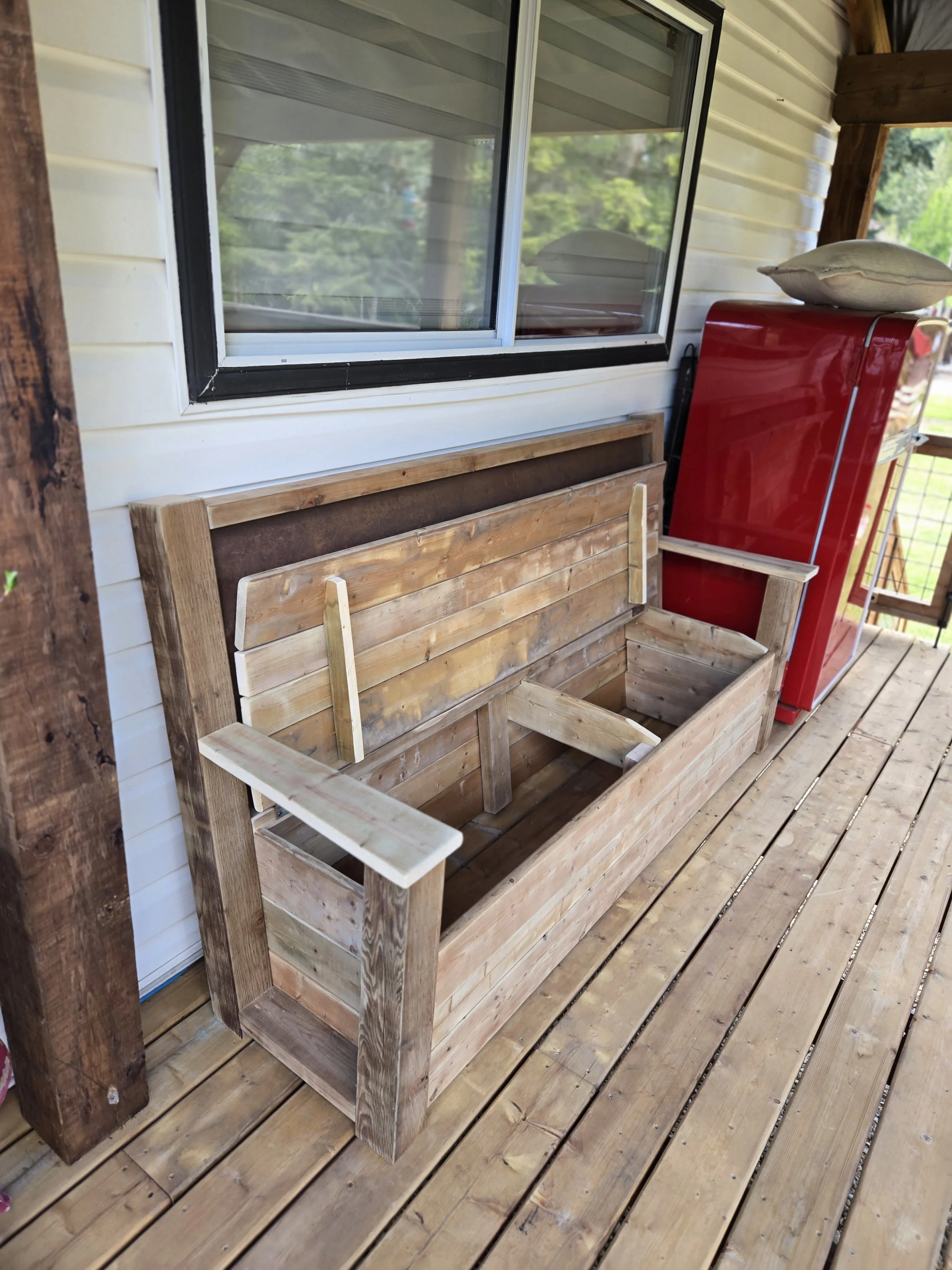 A wooden porch with a large window, a red appliance, a beige pillow on top, and an empty wooden planters box.