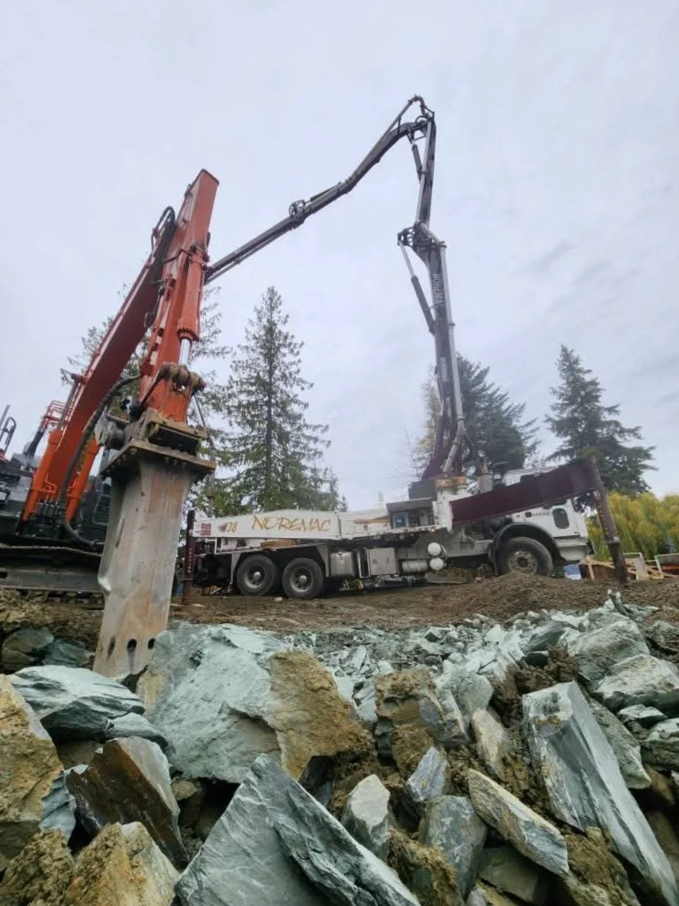 A construction site with a large truck equipped with a hydraulic arm, lifting a concrete pile into place among rocks and dirt, with trees in the background.