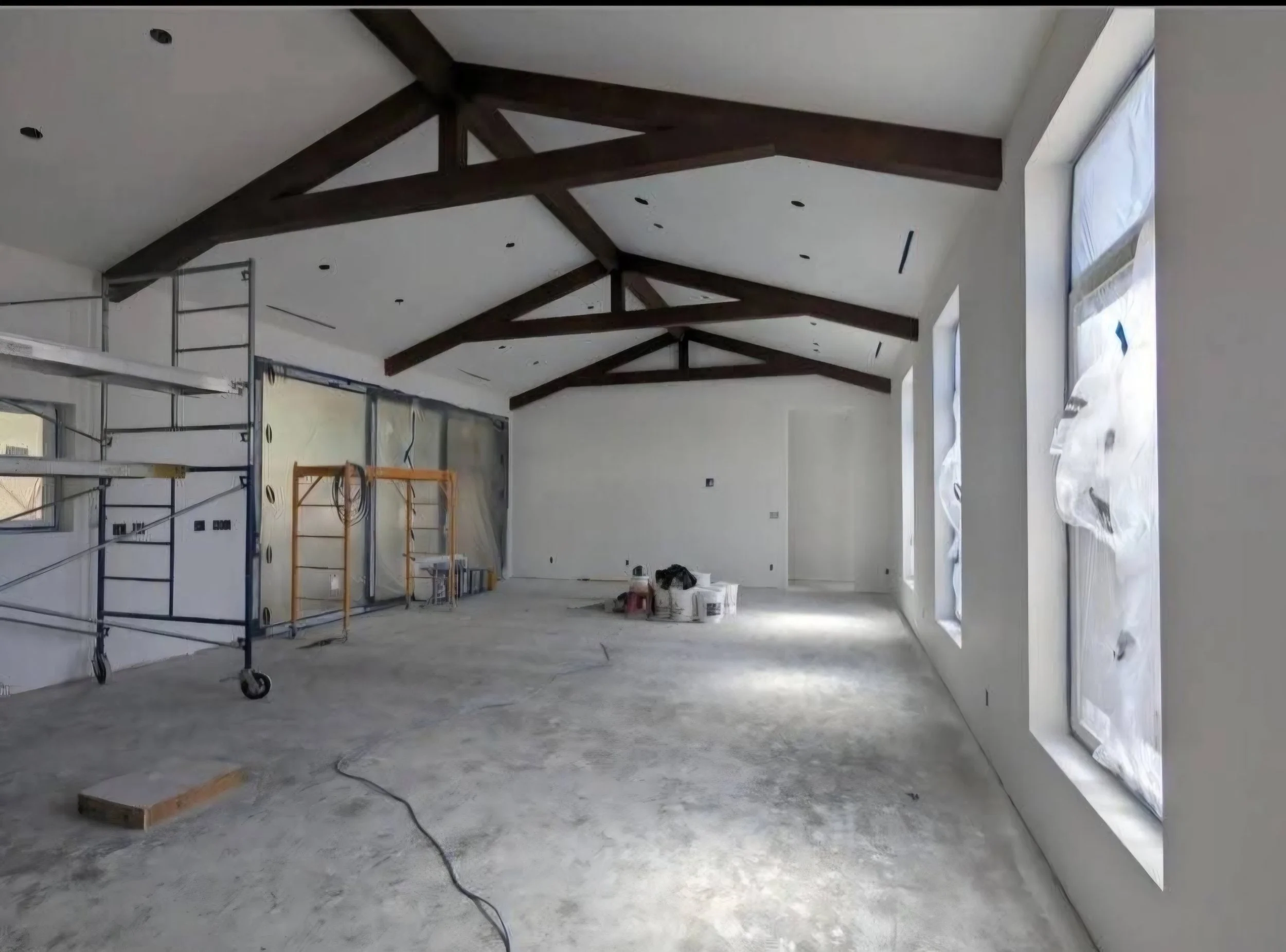 Interior view of a room under construction with exposed wooden beams on the ceiling, large windows covered with protective plastic, and construction tools and materials scattered on the unfinished floor.
