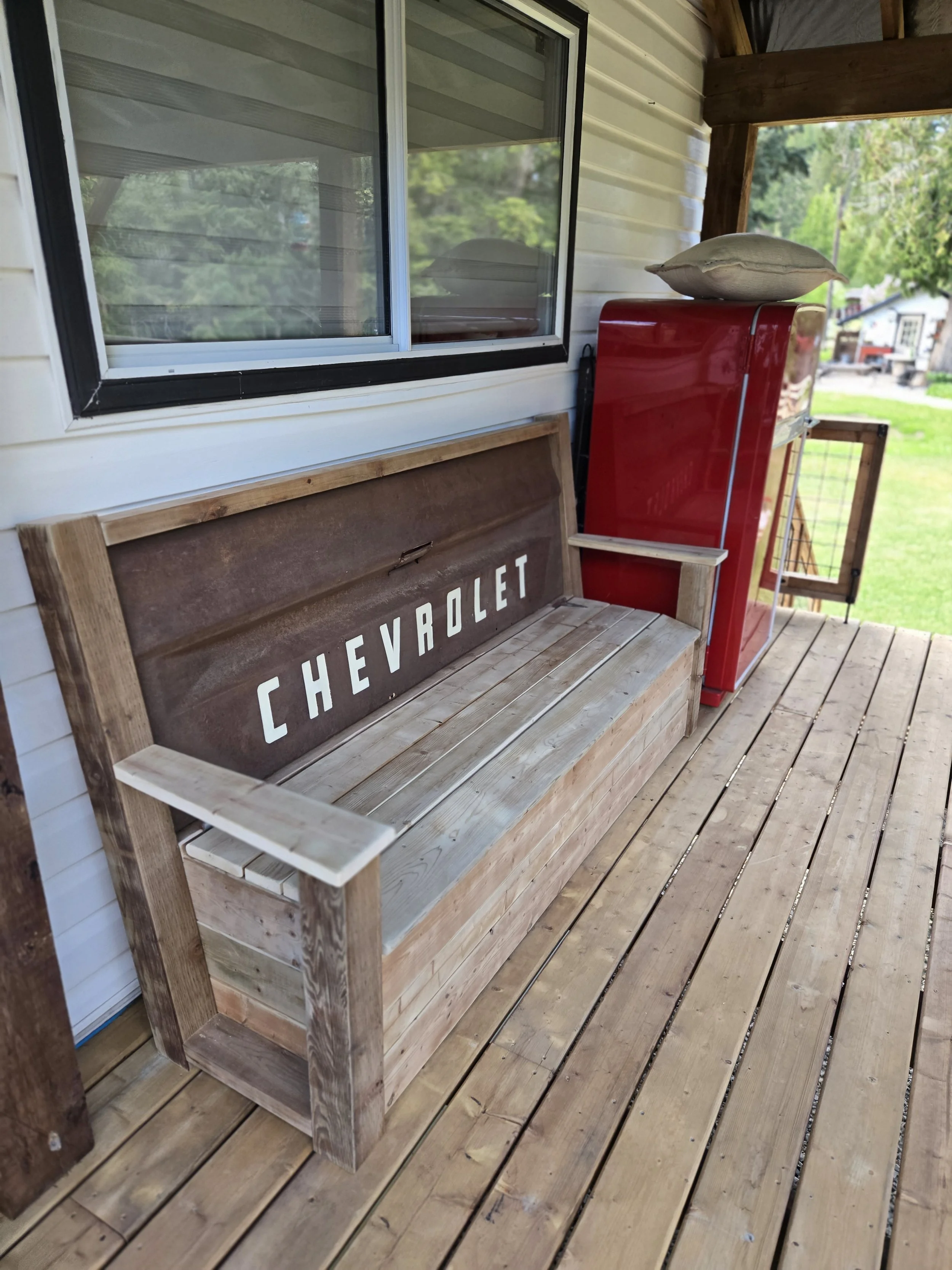 Wooden porch with vintage Chevrolet sign, red vintage fridge, and a pillow on top of the fridge, with green yard and trees in background.