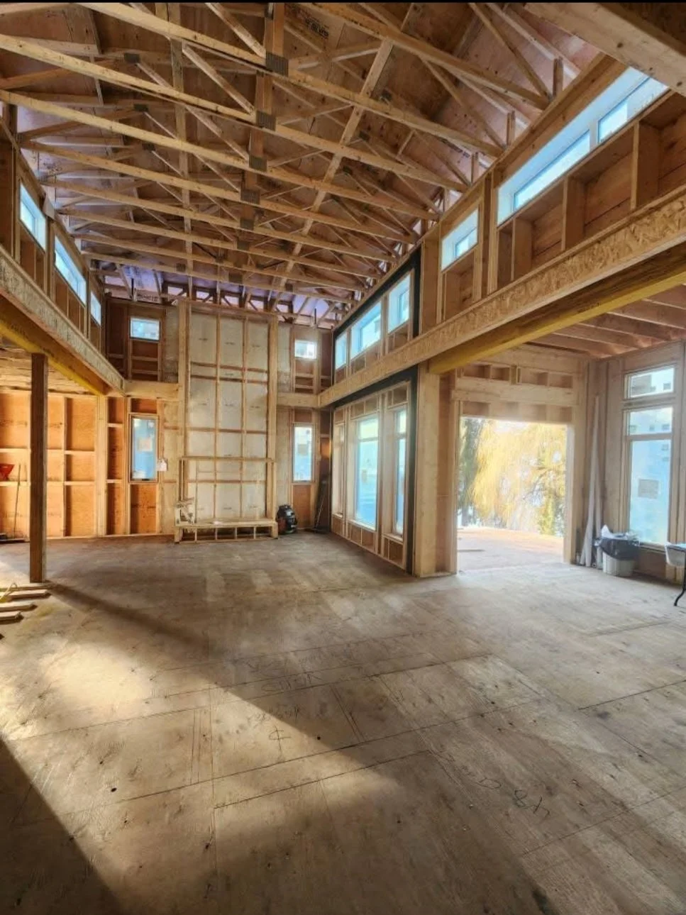 Interior of a house under construction with exposed wooden framing, large windows, and high ceilings.