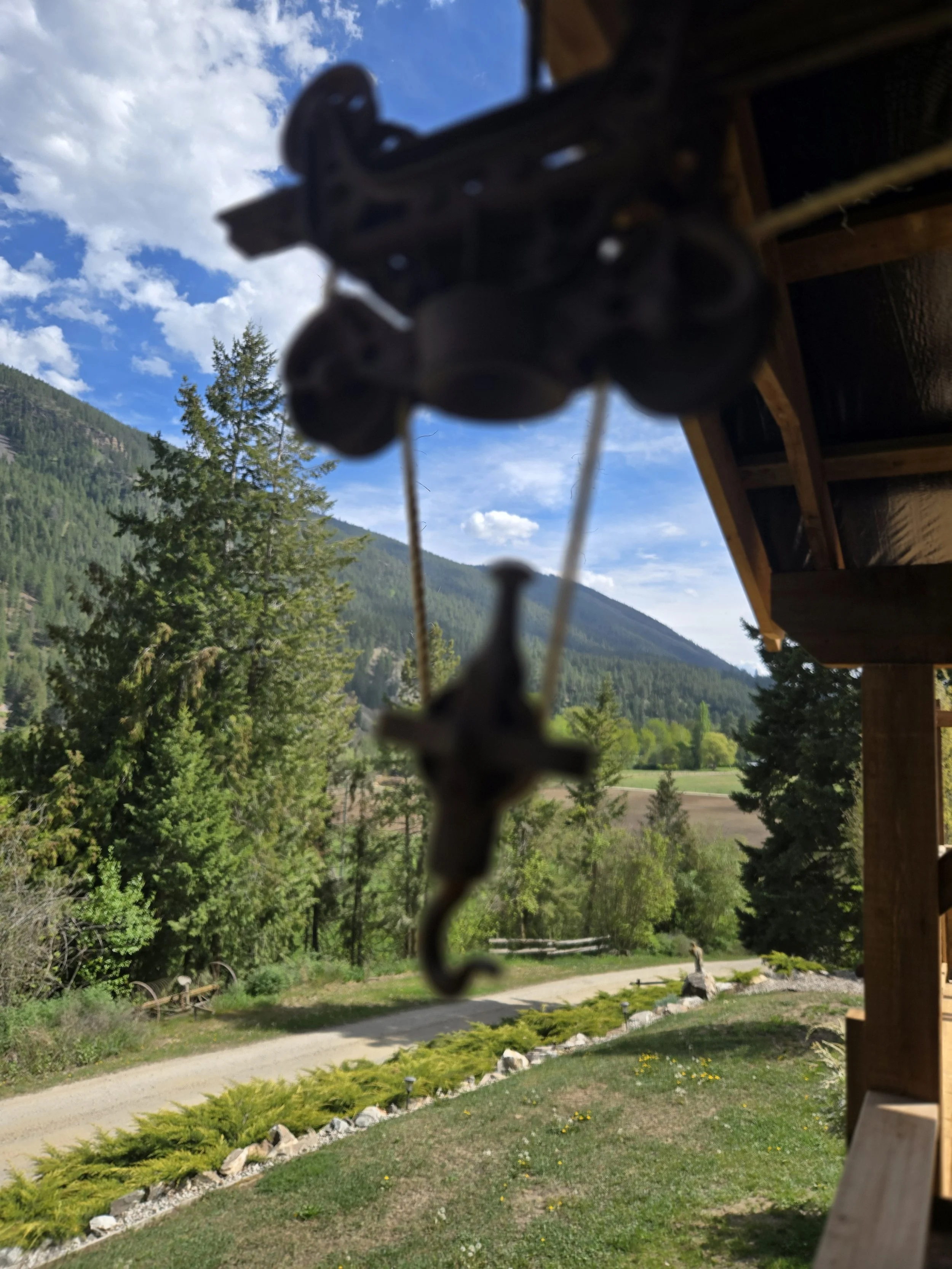 A blurred view of a mountain landscape with evergreen trees, a dirt trail, and a blue sky with white clouds, seen from underneath a wooden porch with a black metal hanging decoration.