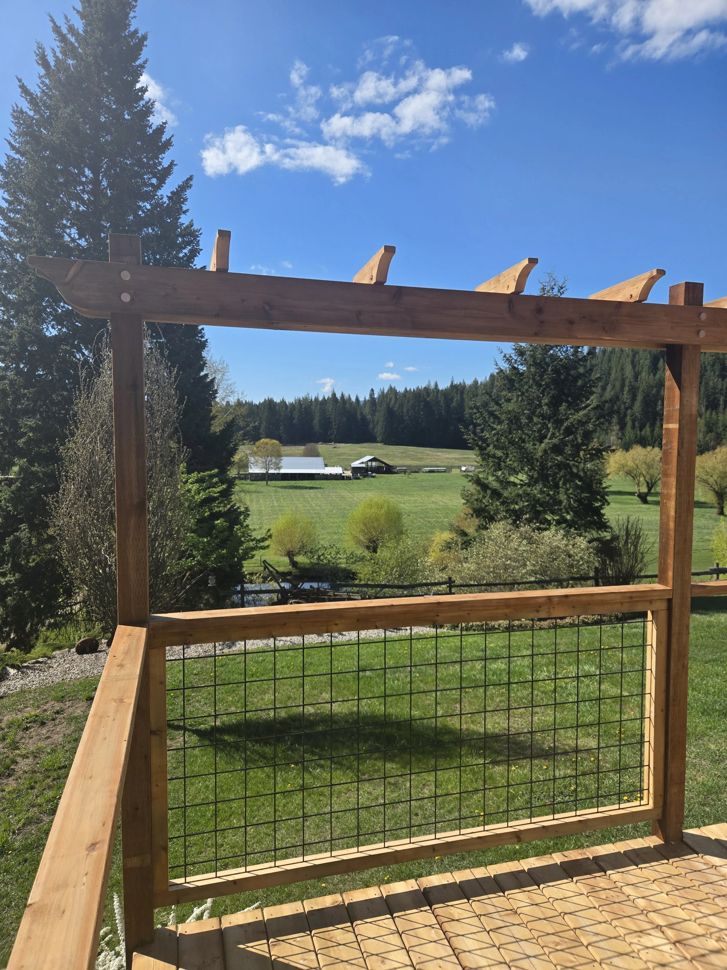 A wooden deck under construction with a view of a countryside landscape featuring green fields, trees, distant farm buildings, and a bright blue sky with a few clouds.
