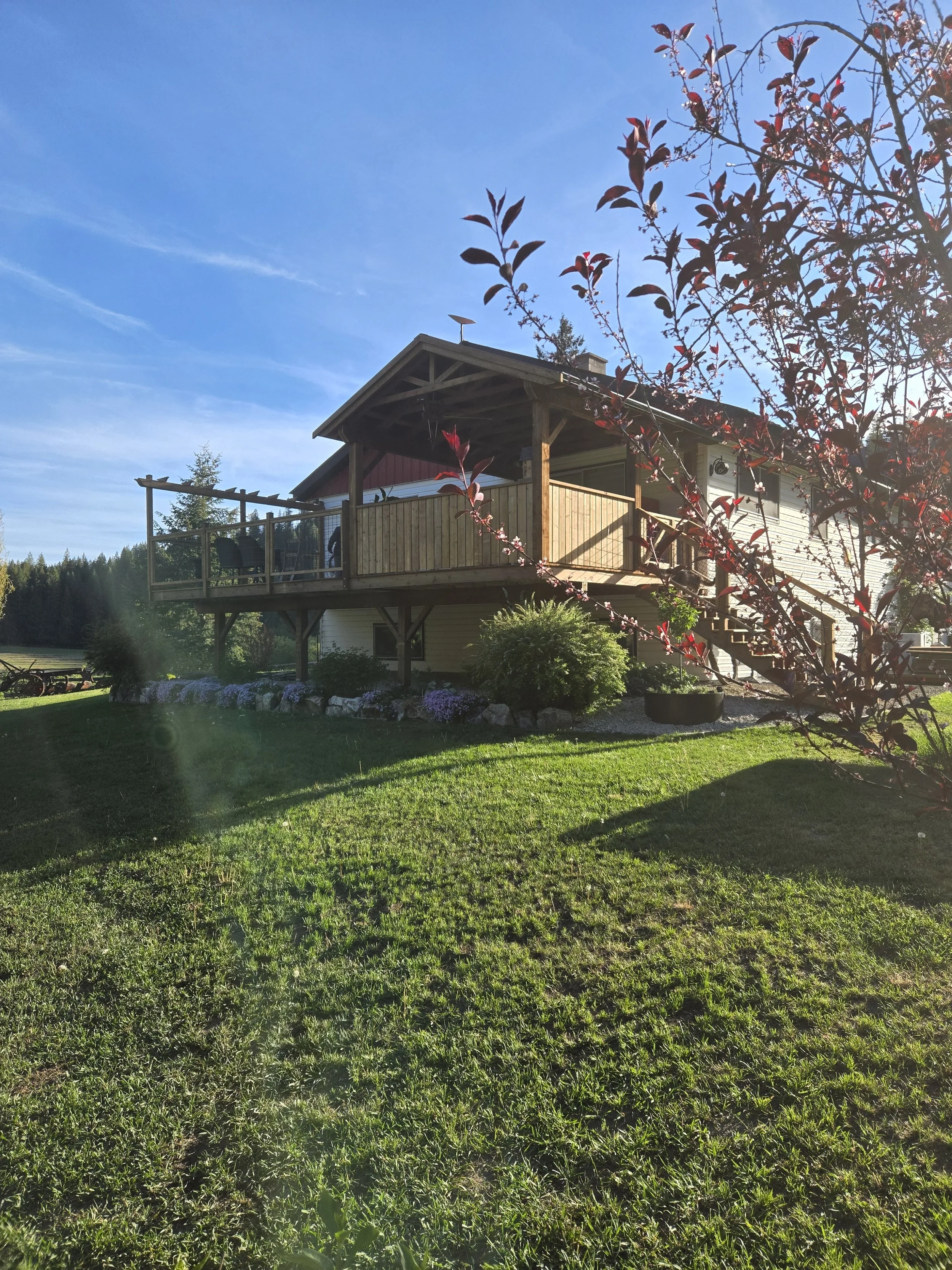 A wooden house with a balcony surrounded by green lawn, trees, and flowering bushes under a blue sky.