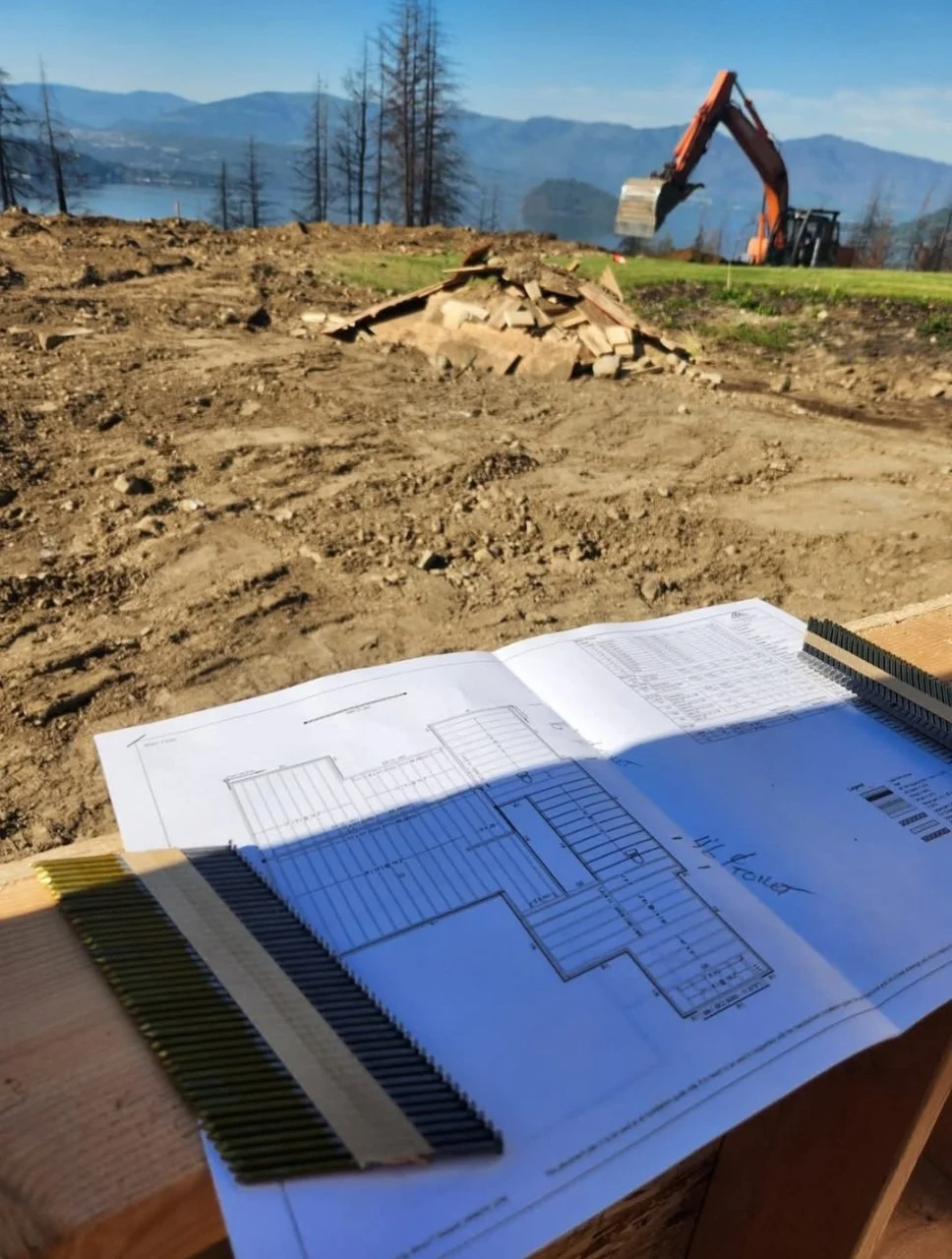 Construction site with a blueprint and samples of building materials on a wooden surface, excavator in the background, dirt and debris in the foreground, and mountains in the distance.
