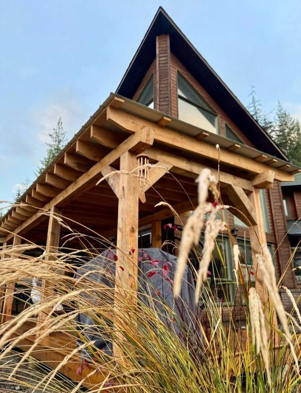 A house with a steep triangular roof and a wooden porch under construction, with tall grass in the foreground.