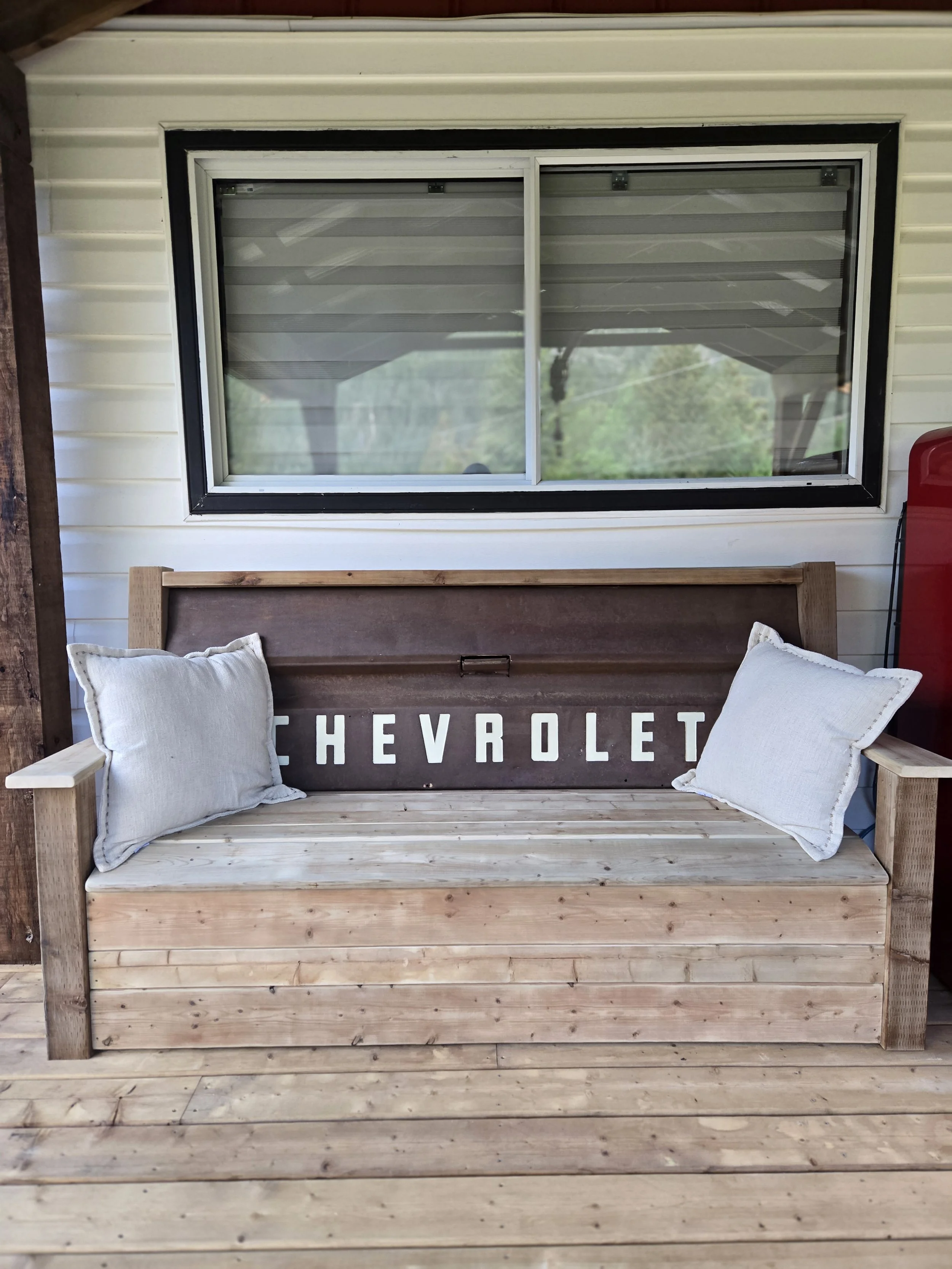 A wooden porch bench with white cushions placed on each side in front of a window, and a sign behind it with the word "CHEVROLET".