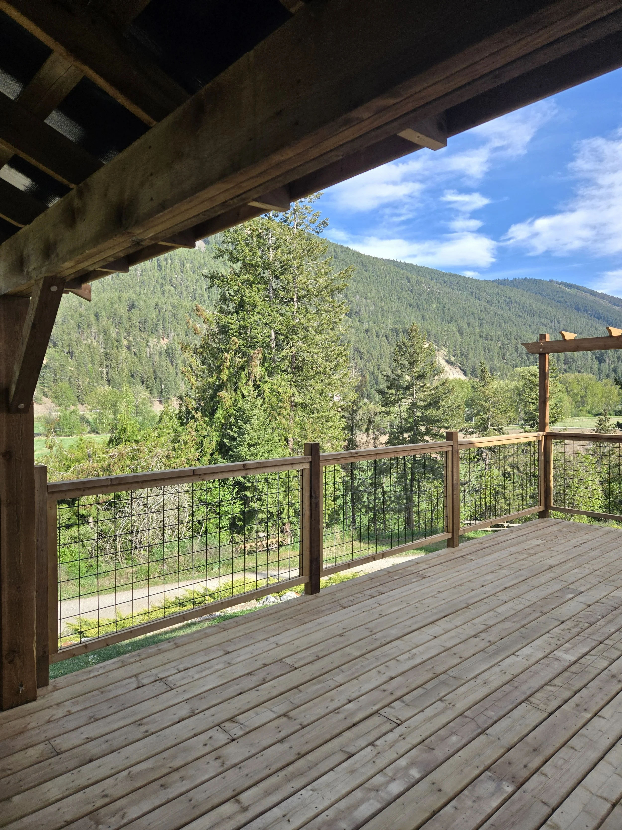 Wooden deck with a railing overlooking a green forested mountain landscape under a partly cloudy blue sky.
