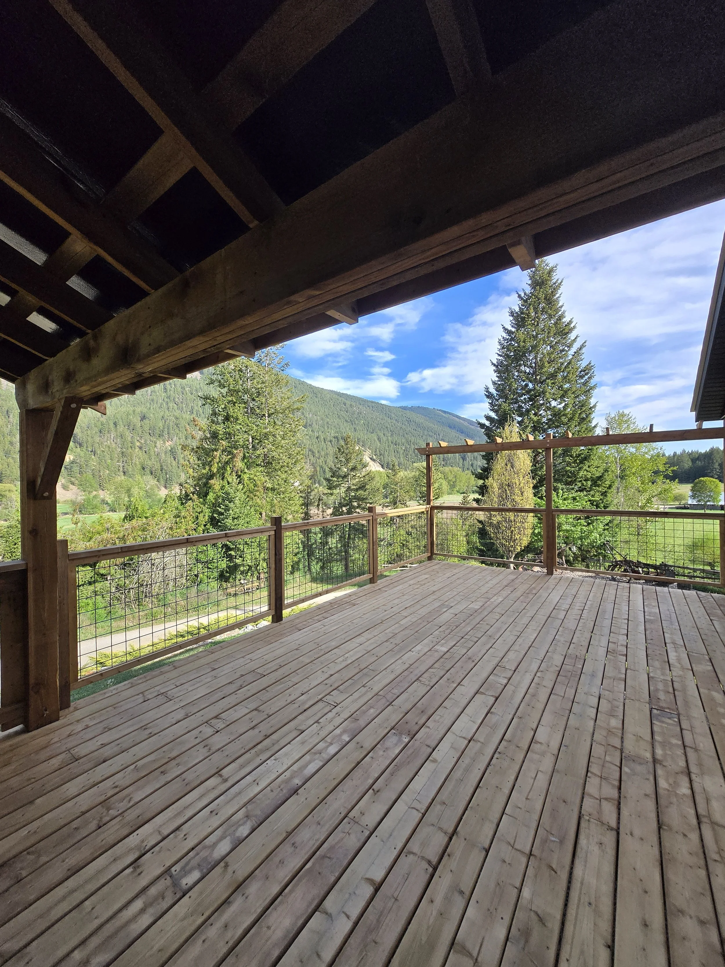 View from a wooden porch with railings, overlooking a lush green landscape with trees, mountains, and a partly cloudy blue sky.