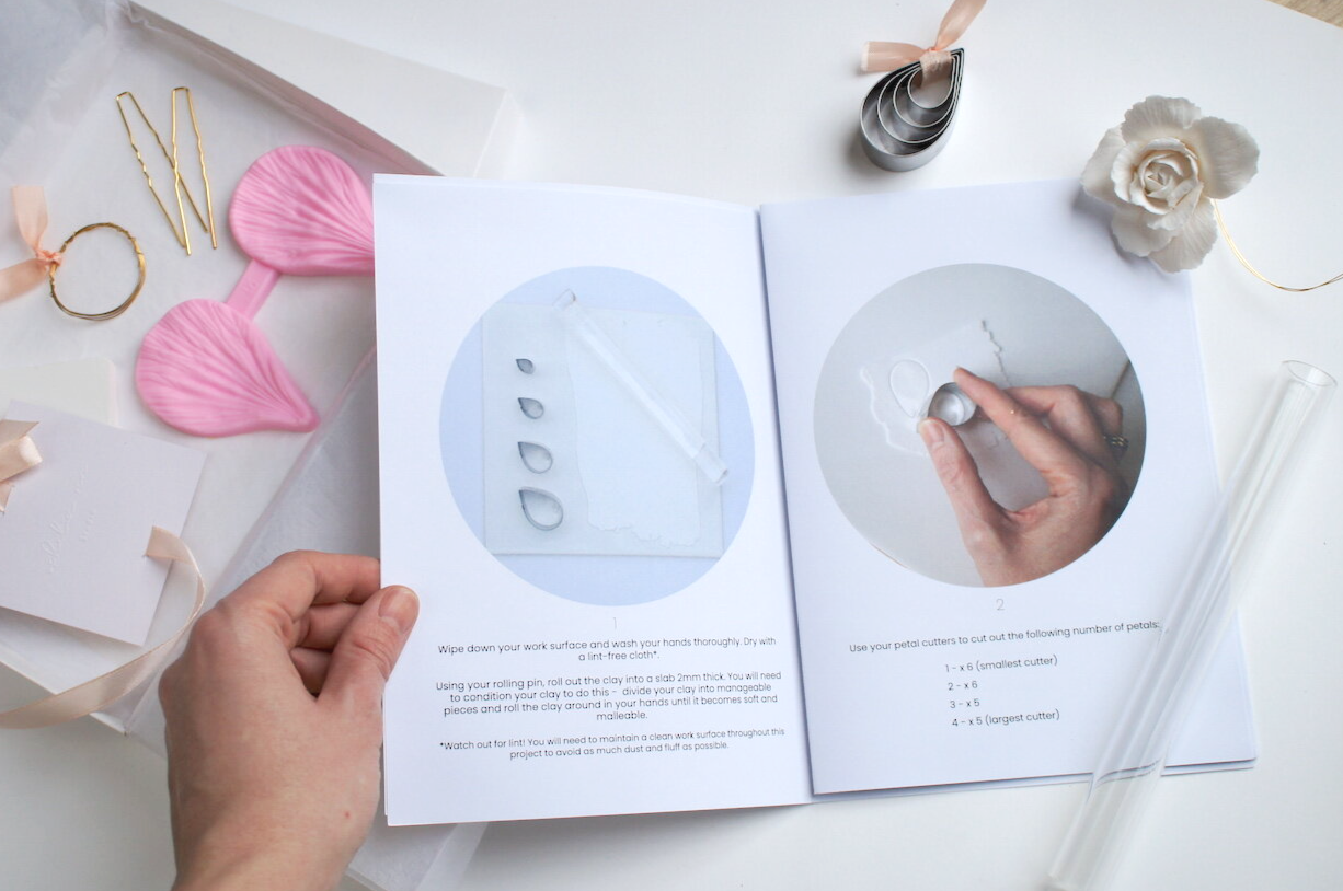 Open craft instructions manual surrounded by craft supplies, including pink petal-shaped utensils, gold hairpins, petal cutters, and a white rose flower. All on a white surface.