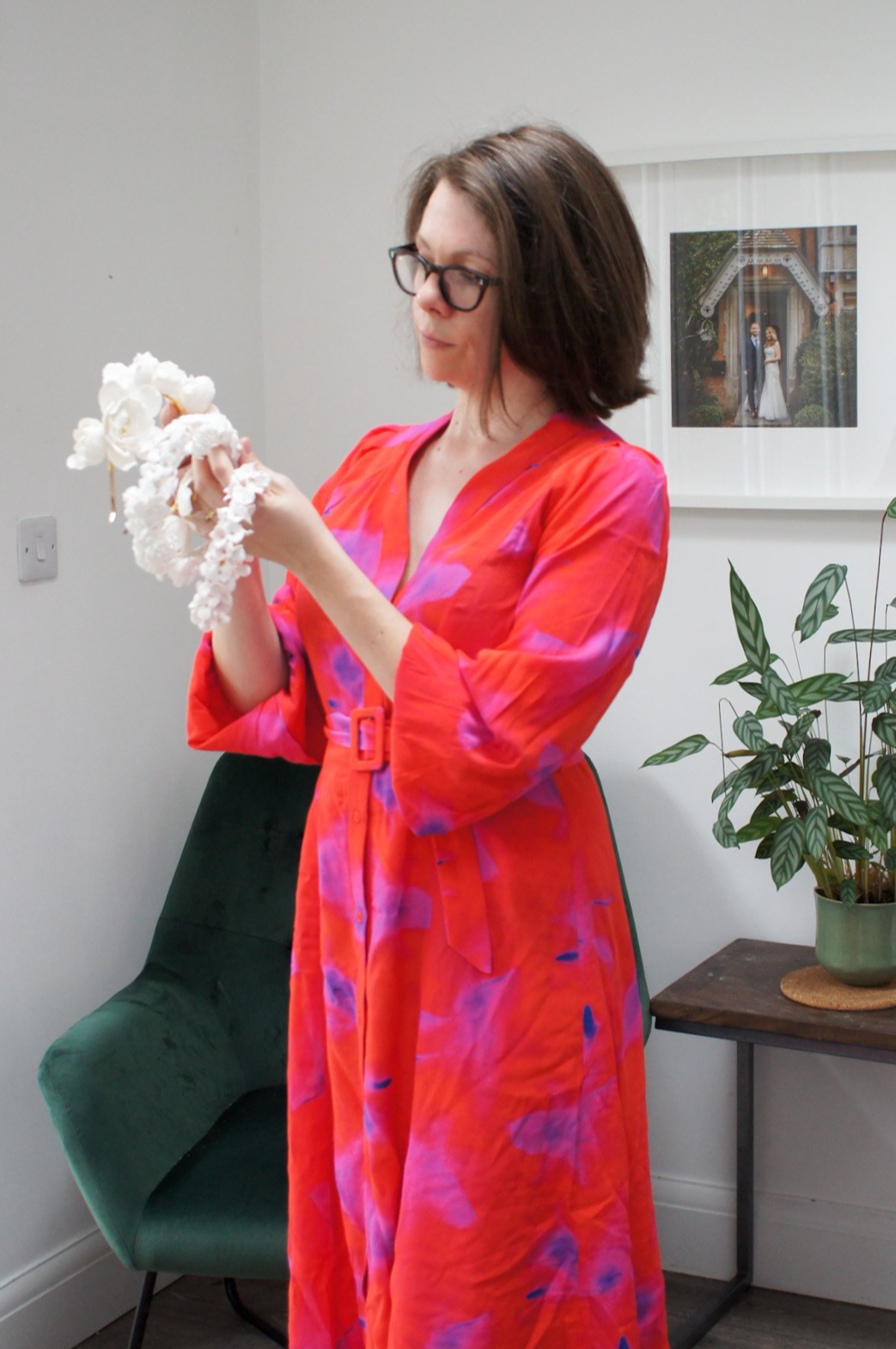 A woman with shoulder-length brown hair and glasses wearing a colorful red and purple dress standing in a room with white walls, holding white handmade floral bridal headbands and crowns