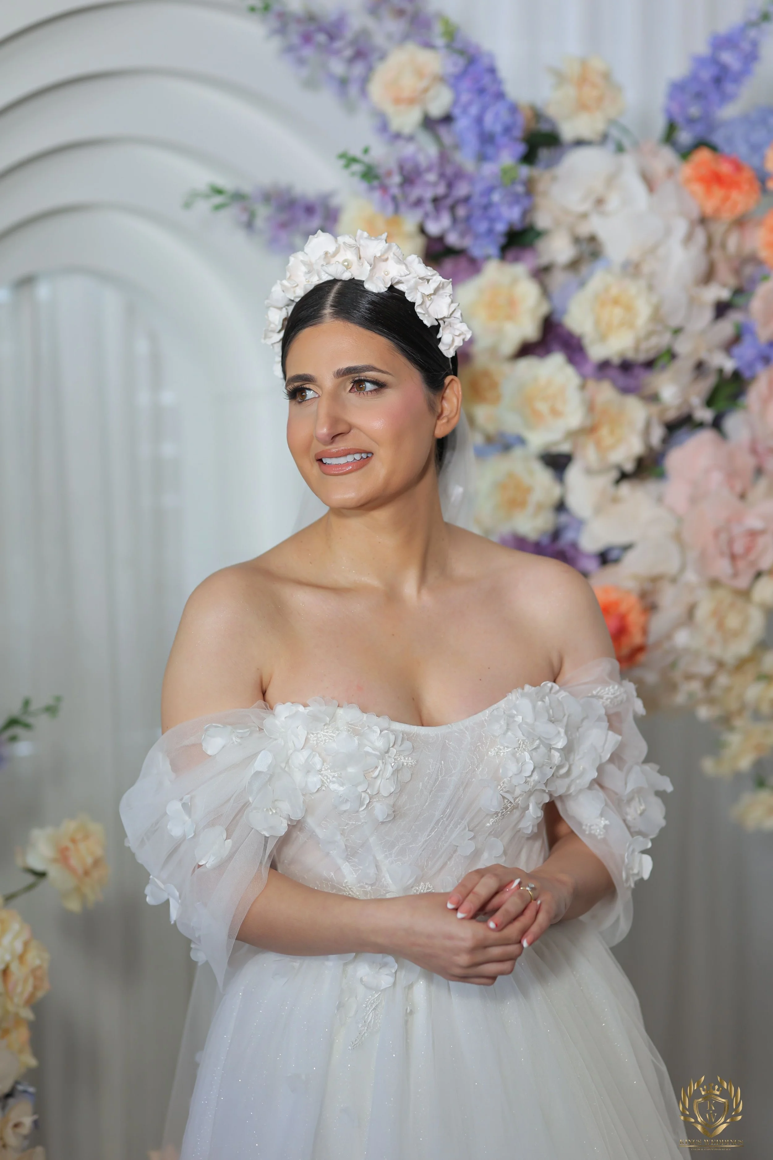 A bride wearing a white wedding dress with floral embellishments, a floral headpiece, and a veil, standing in front of a large bouquet of pastel-colored flowers and a white decorative backdrop.