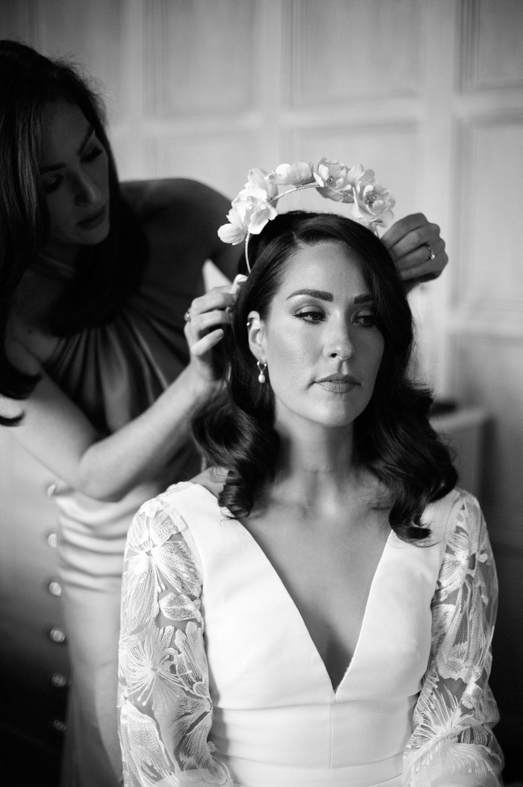 A woman with dark, wavy hair is sitting with a floral crown being placed on her head by another woman. The scene is in black and white, and the woman has a serene expression, wearing a white dress with lace sleeves.