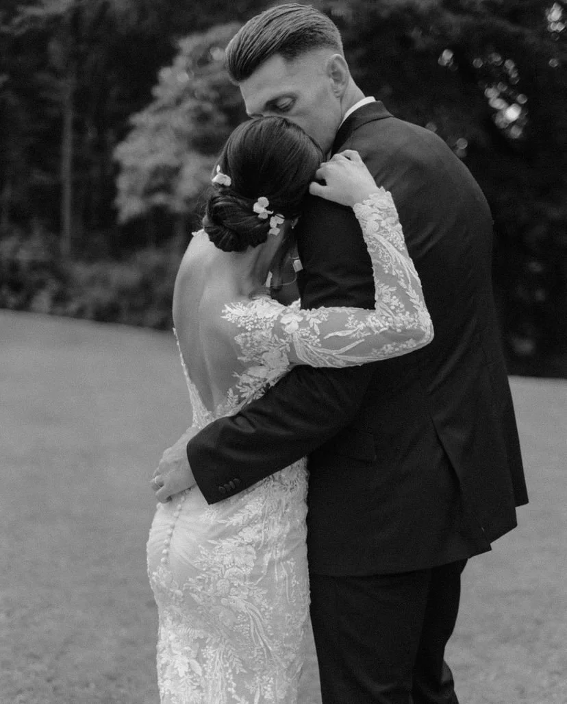 A black and white photo of a couple embracing outdoors, with the woman wearing a lace wedding dress and white floral hairpins in her hair. The man is in a suit, sharing an emotional kiss.