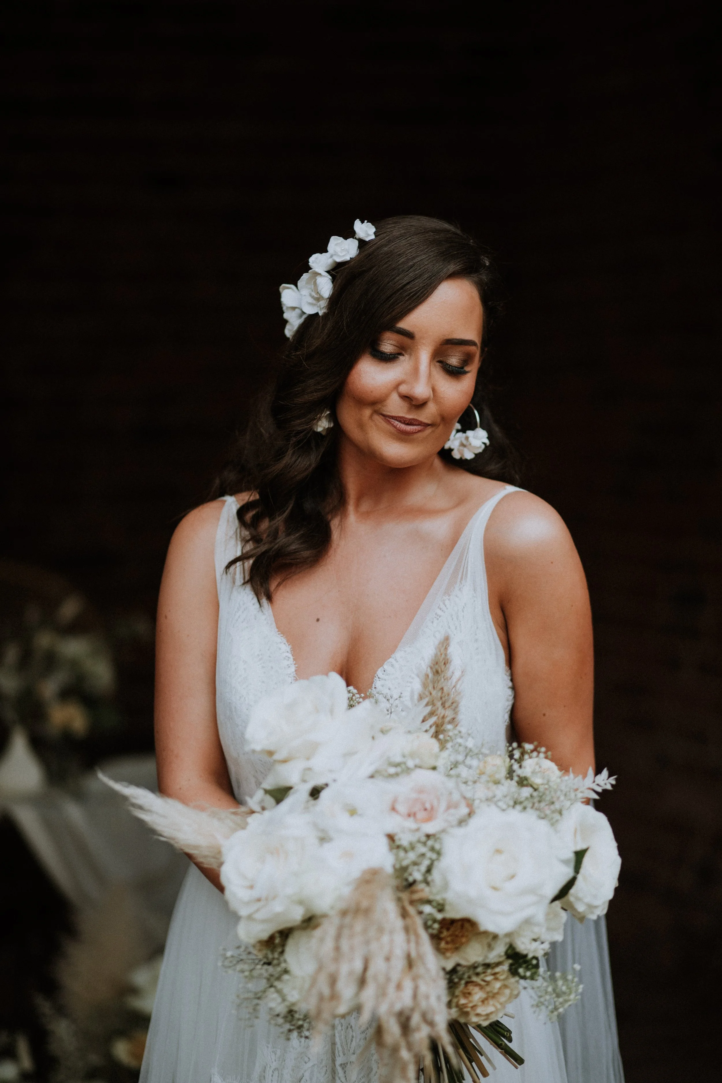 A bride with dark hair styled in loose waves, wearing a white lace wedding dress with floral accessories in her hair and matching earrings, holding a bouquet of white and blush flowers, on a dark background.