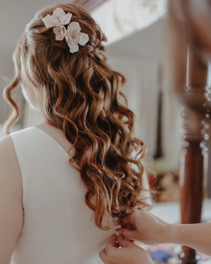 A woman with long, curly red hair styled with white flowers, wearing a sleeveless white dress.