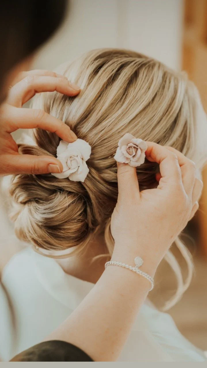 A woman with blonde hair is having her hair styled into an updo with two pink clay handmade flower hairpins being placed in her hair by two hands.