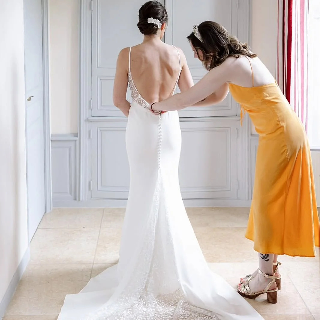 A bride in a form-fitting white wedding gown and white bridal hairpins to match is being assisted with final preparations by a woman in a yellow dress.