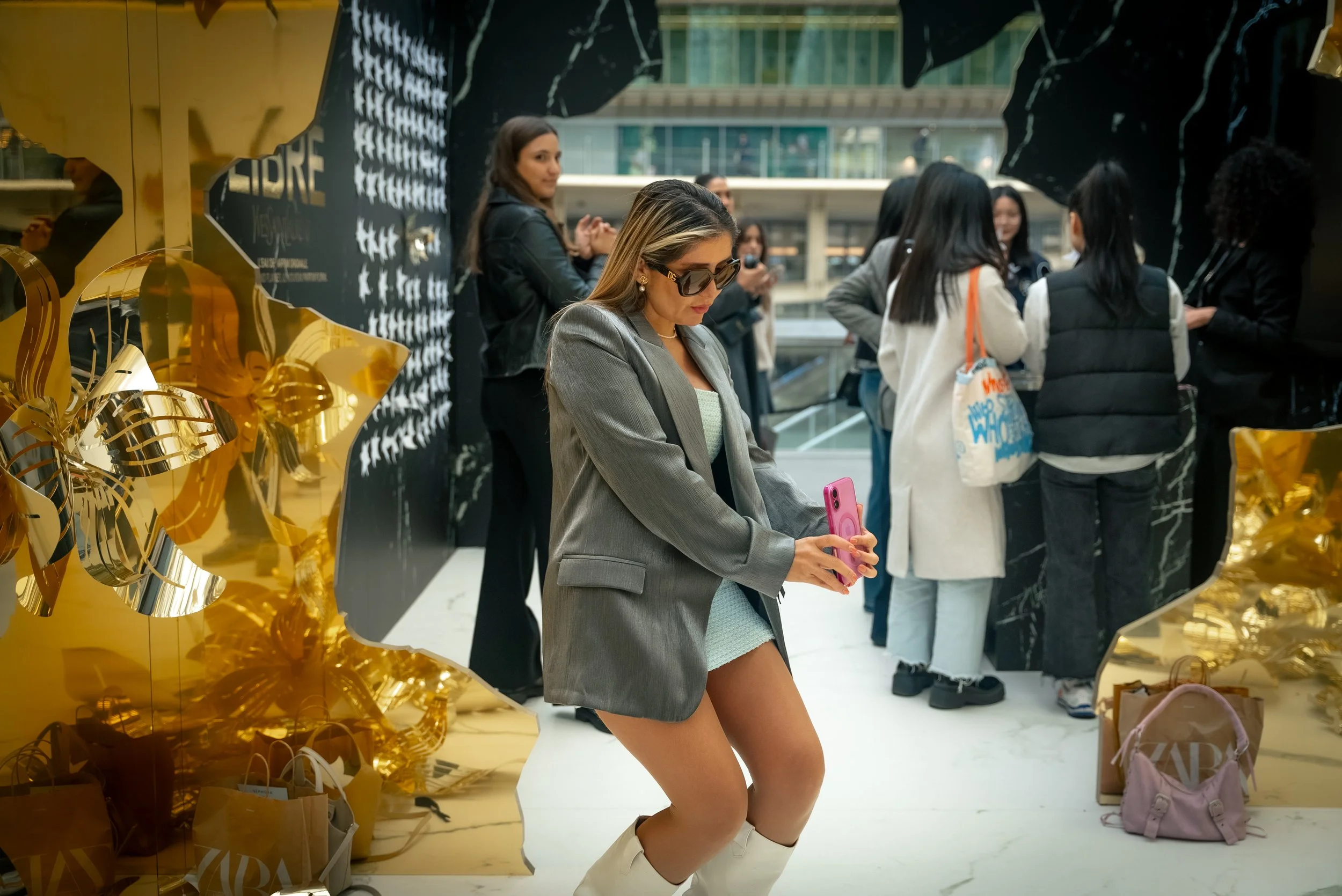 Une femme en blazer gris, portant des glasses de soleil et des bottes blanches, posant pour une photo avec un téléphone rose devant un mur décoré de ballons et d'une grande décoration noire et or, entourée de plusieurs autres femmes parlant en groupe