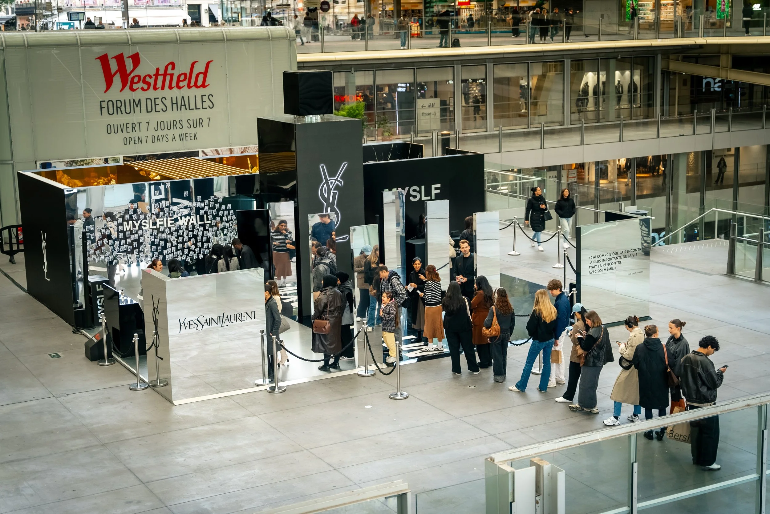 Une file d'attente de personnes devant un stand de la marque Yves Saint Laurent à l'intérieur d'un centre commercial.
