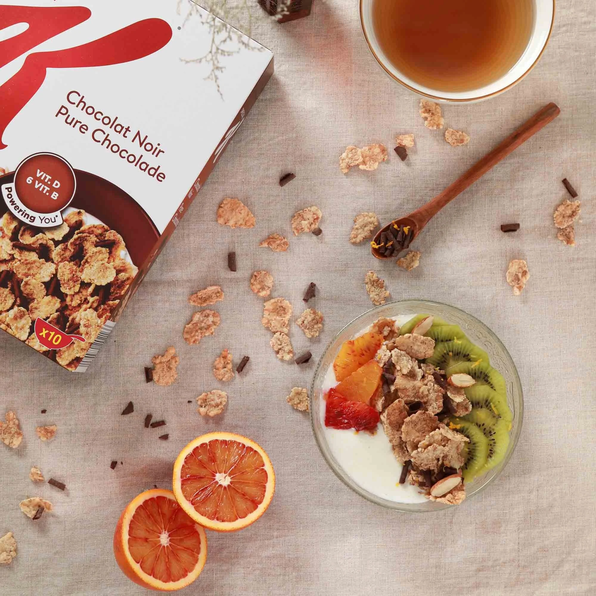 Bol de yaourt garni de tranches de kiwi, orange et autres fruits, avec des céréales et des amandes, à côté d'une moitié d'orange et d'une boîte de céréales au chocolat